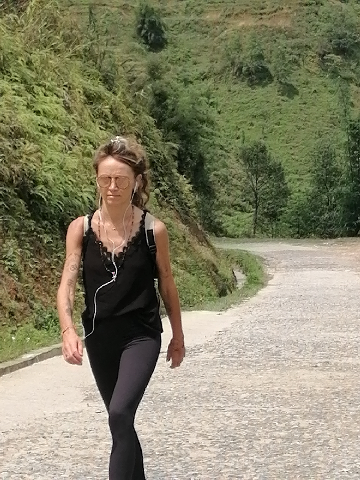 A woman is walking on a dirt road. She wears a black tank top with lace trim, black leggings, and tan-tinted sunglasses. She has a backpack and earphones. The road curves and has vegetation on both sides. In the background is a hillside covered with trees and bushes. The location is Sa Pa Ward, Vietnam.