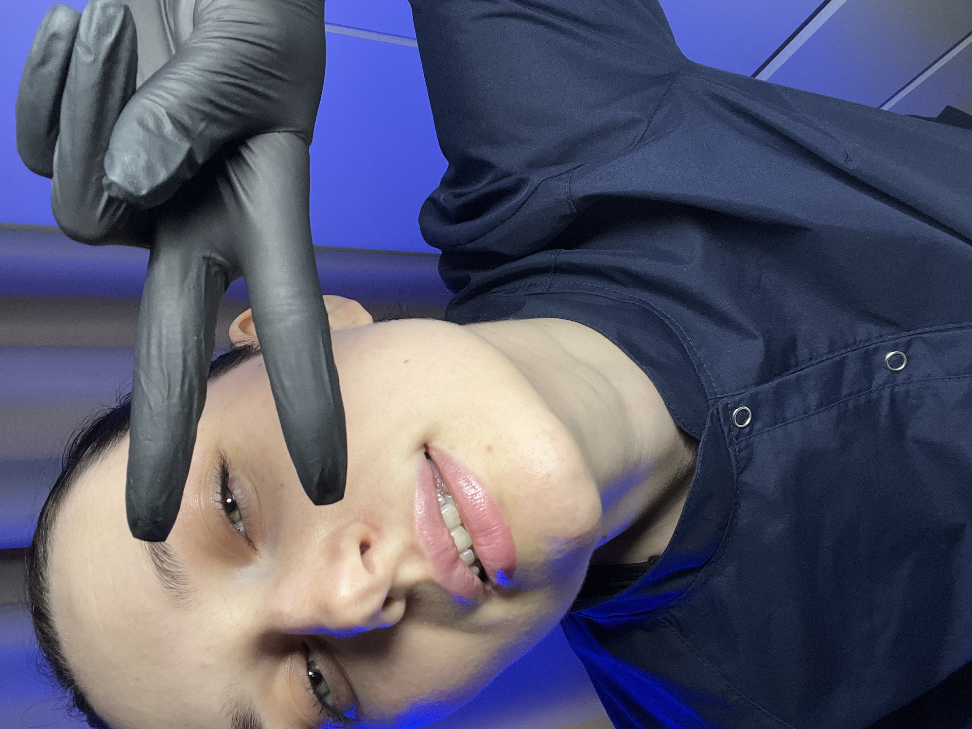 An image captured in Tyumen, Russia, depicts a female subject from the chest up, centered in the frame. The subject possesses fair skin, dark hair pulled back, and appears to be a young adult. She wears a dark navy blue uniform-style top with a stand-up collar and silver snap fasteners on the front placket. Her right hand, encased in a black nitrile or latex glove, is raised towards her face, forming a "V" sign with the index and middle fingers extended and separated. This gesture is positioned above her right eye, slightly obscuring the upper portion of her forehead and eyebrow. Her eyes are open and directed towards the viewer, and her lips are slightly parted, revealing upper teeth, with a visible sheen, possibly from gloss or moisturizer. The background is illuminated with a strong, uniform blue light, displaying subtle geometric lines or paneling towards the right side of the frame. The blue illumination casts a noticeable hue onto the subject's skin and lips.