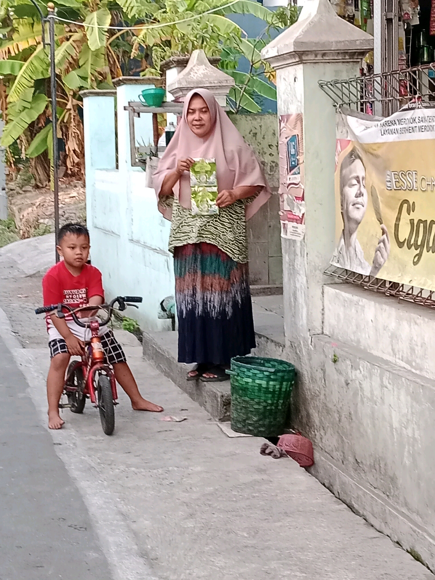 In Waru, Indonesia, a young boy in a red shirt and checkered shorts sits on a small red bicycle, facing left. He is barefoot and appears to be on a sidewalk or pathway. Beside him, to his right, stands an adult woman wearing a pink hijab and a patterned skirt. She is holding a green package. Behind the woman, a poster is visible on a wall, depicting a man's face. The environment includes a wall, a metal fence, a green woven basket, and lush green foliage, suggesting an outdoor setting with vegetation. The ground is paved, with a street visible to the left of the boy.