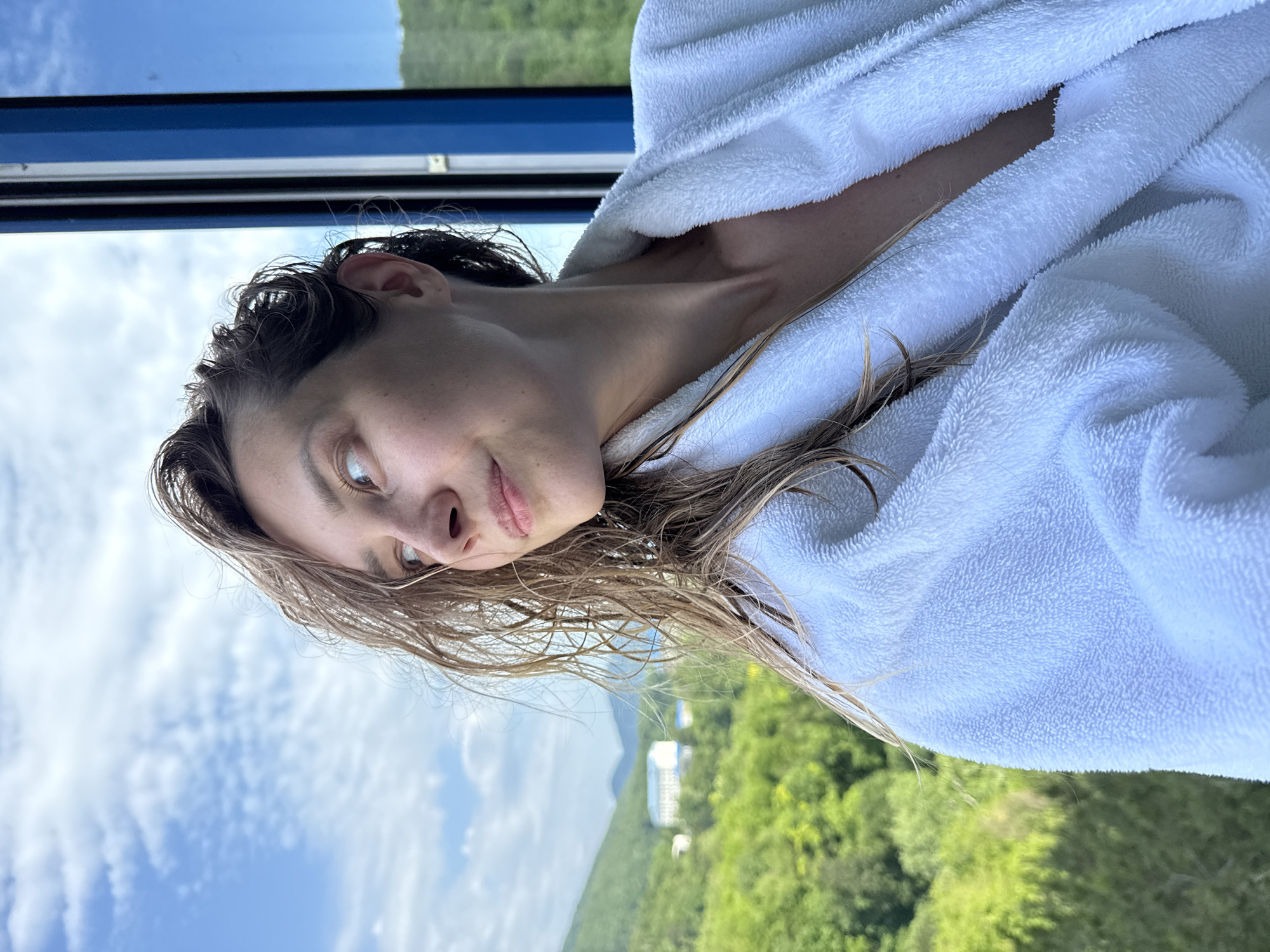A medium shot captures a woman from the chest up, facing left and looking out a window. She is wearing a fluffy white bathrobe, which is open at the neck, revealing a glimpse of her collarbone and upper chest. Her hair is damp and appears to be a light brown, with strands falling forward and to the side. The woman has a light complexion and is looking towards the left side of the frame.
The background offers a view of a lush green landscape with rolling hills covered in dense trees. In the distance, a white building with a blue roof is visible, situated among the greenery. The sky above is a bright blue with scattered white clouds. To the right of the woman, a portion of a blue window frame is visible, suggesting she is indoors looking out. The overall impression is a serene moment captured with a scenic backdrop. The location is identified as Nebug, Russia.