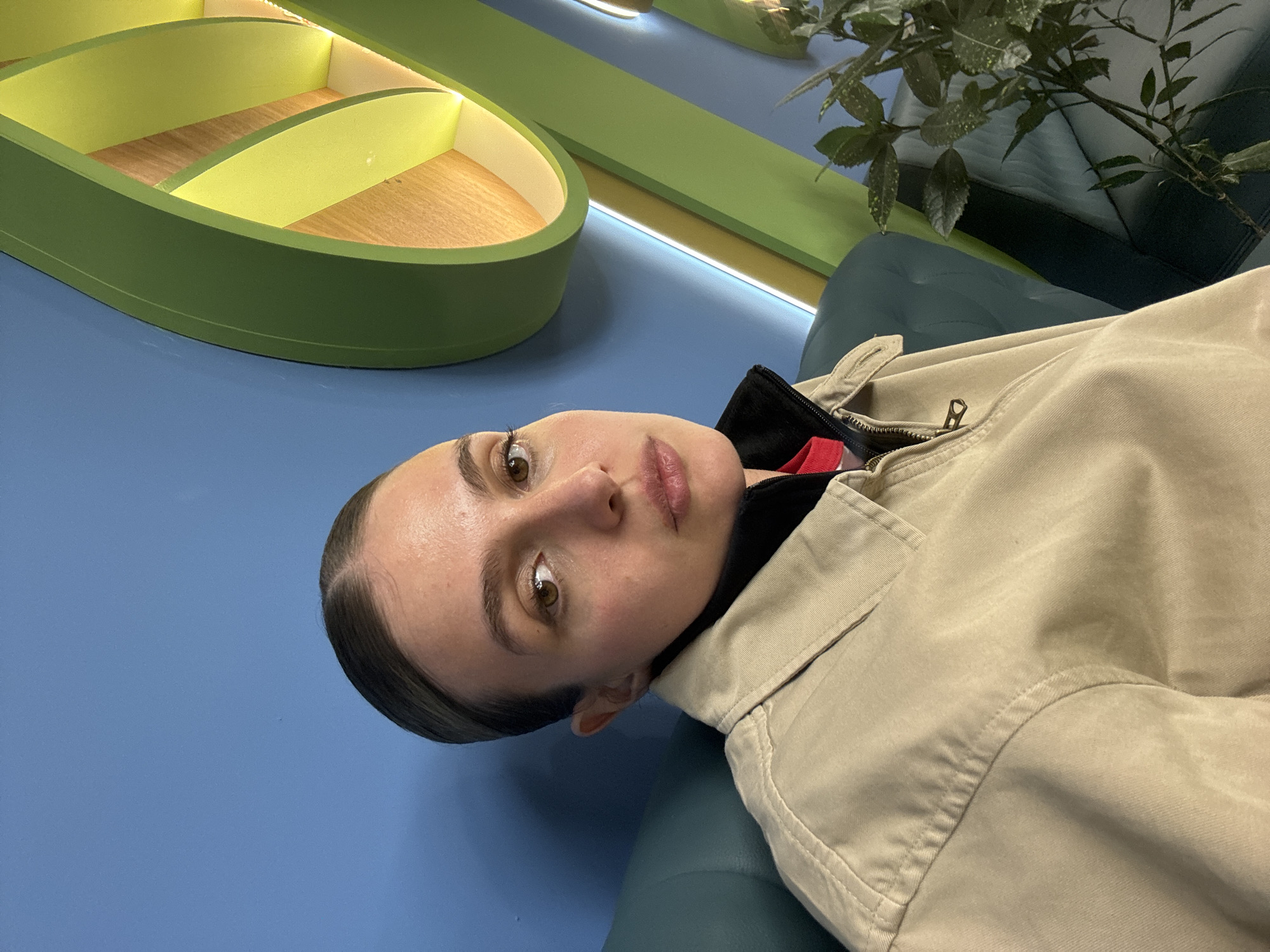 A medium shot shows a woman with dark hair slicked back, wearing a tan jacket, seated in a green chair against a blue wall. Behind her is green decor, including a shelf-like structure with wooden shelves and a leafy plant. The location is likely an interior space in Tbilisi, Georgia, given the decor and clothing style.