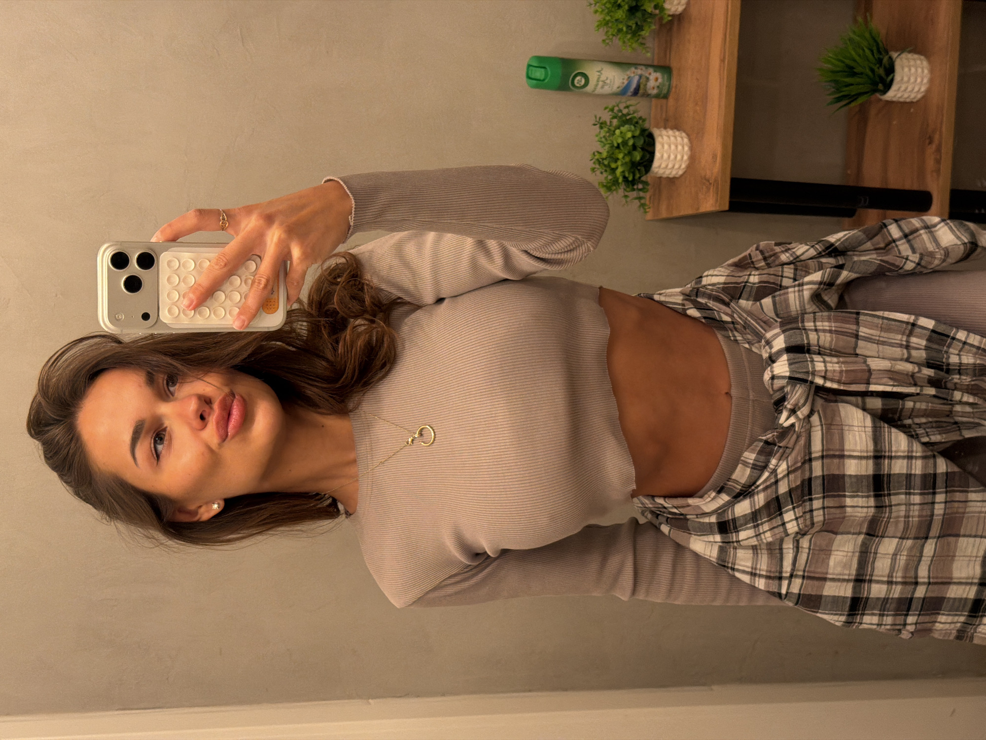 In a dimly lit indoor space, possibly a fitting room or bathroom, a young woman stands facing a mirror, taking a selfie with her phone. She has long, brown hair and is wearing a light gray, long-sleeved crop top, revealing her midriff. A plaid shirt is tied around her waist. She is accessorized with a delicate gold necklace and a ring on her right hand. Behind her, a shelf displays small potted plants and an air freshener. The wall is a neutral gray. There is no indication of the city of Krasnoyarsk, Russia.