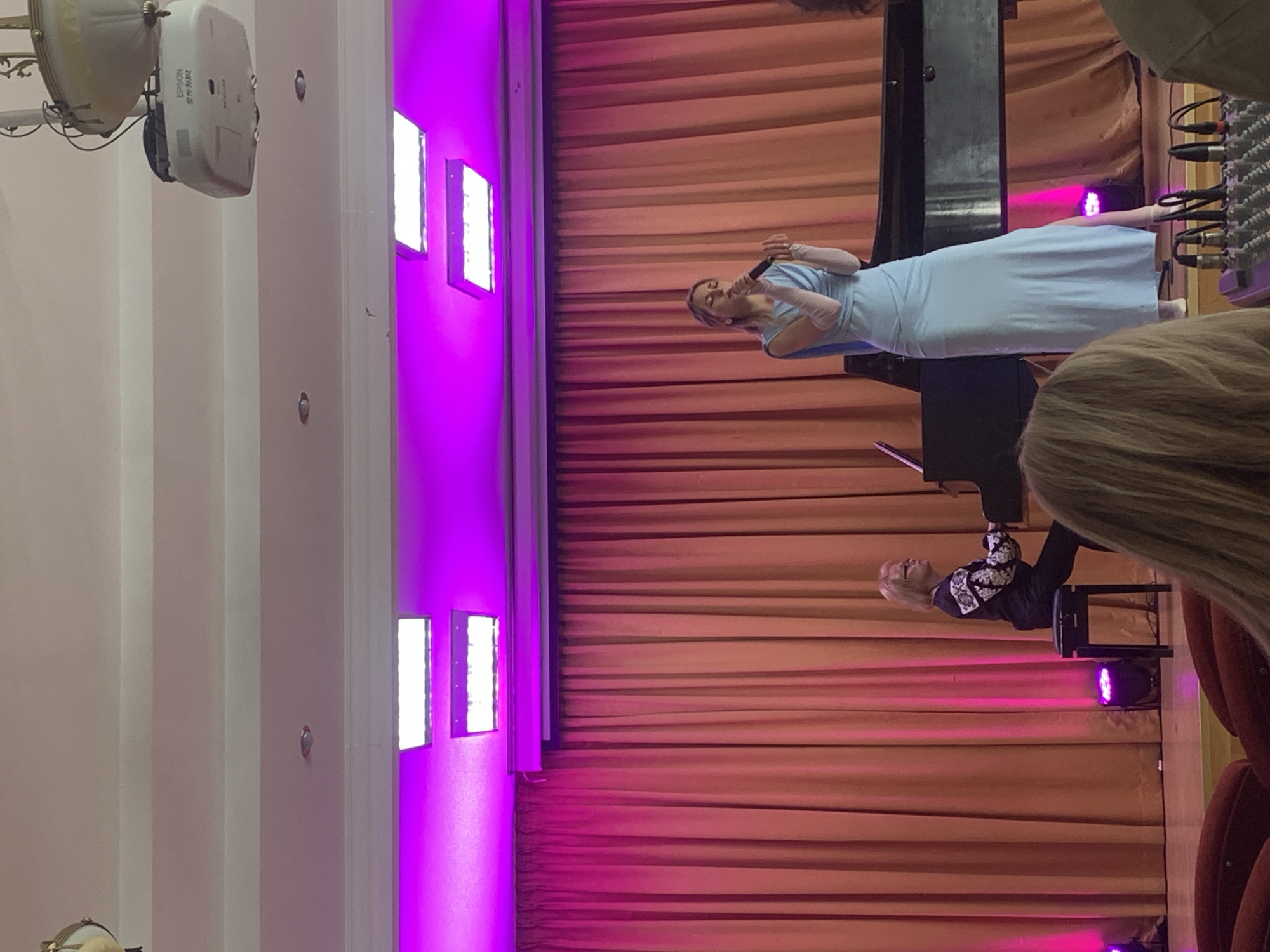 This image captures a performance on a stage illuminated by pink and purple lights. A woman in a light blue, floor-length gown is singing into a microphone, her left hand extended. To her left, an older woman with blonde hair and glasses plays a black grand piano, seated on a stool. Behind them, a large, rust-colored curtain serves as the backdrop. The stage floor appears to be light-colored. In the foreground, the tops of red seats are visible, suggesting an audience is present. A mixing board with numerous cables is also visible in the lower right corner of the frame. The venue is not identified, and there is no information about the location being in the city of Kyiv, Ukraine.