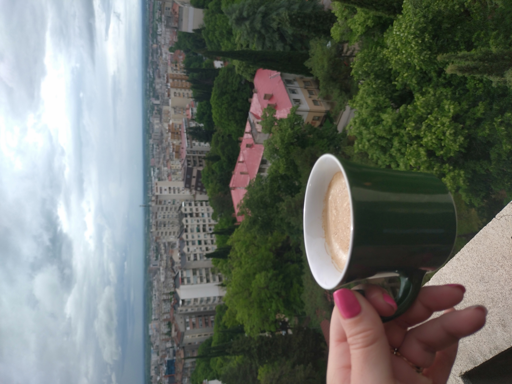 A medium, wide shot captures a woman's hand holding a green and white coffee mug, superimposed over an urban landscape of Stara Zagora, Bulgaria. The foreground features the woman's hand, holding the mug with a light brown beverage. The woman's nails are painted a bright pink. Behind this, the city of Stara Zagora is visible, featuring a mix of buildings and greenery. Some of the buildings have red roofs. The sky is overcast with a layer of gray clouds. The perspective is from an elevated position, likely a balcony or high-rise. The image orientation is vertical.