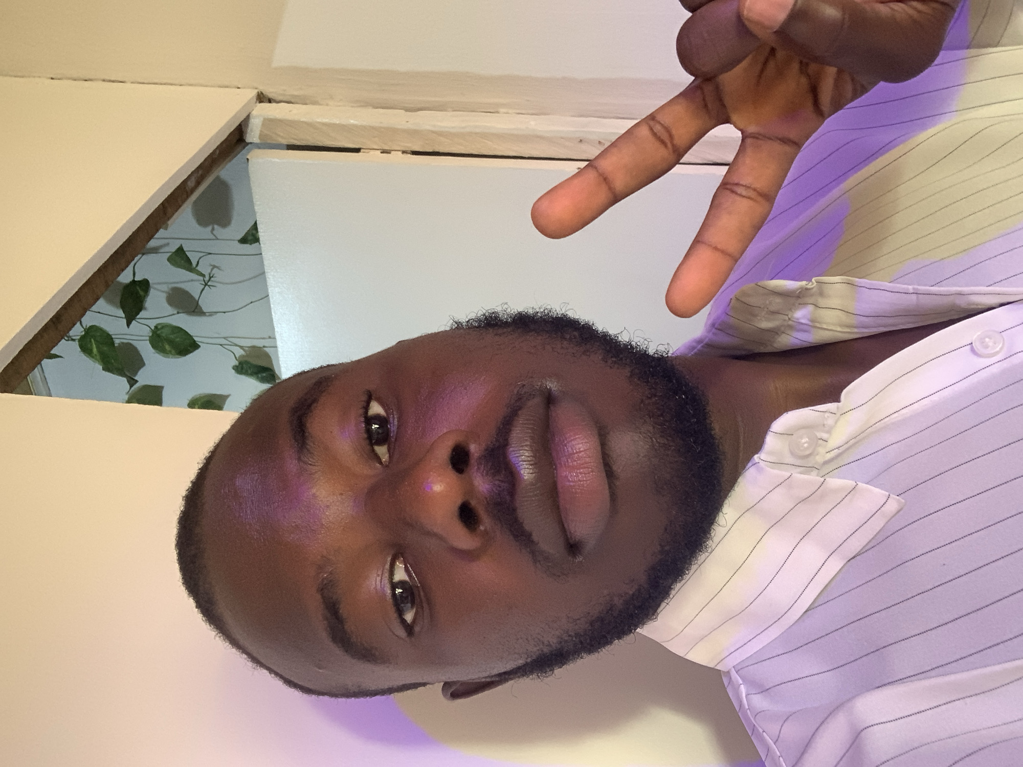 A dark-skinned man with a beard is posing for a selfie, making a peace sign with his right hand. He is wearing a light-colored, vertically striped collared shirt. The background shows a light-colored wall and a doorway with a glimpse of a leafy decorative element. Location cannot be determined based on the image.