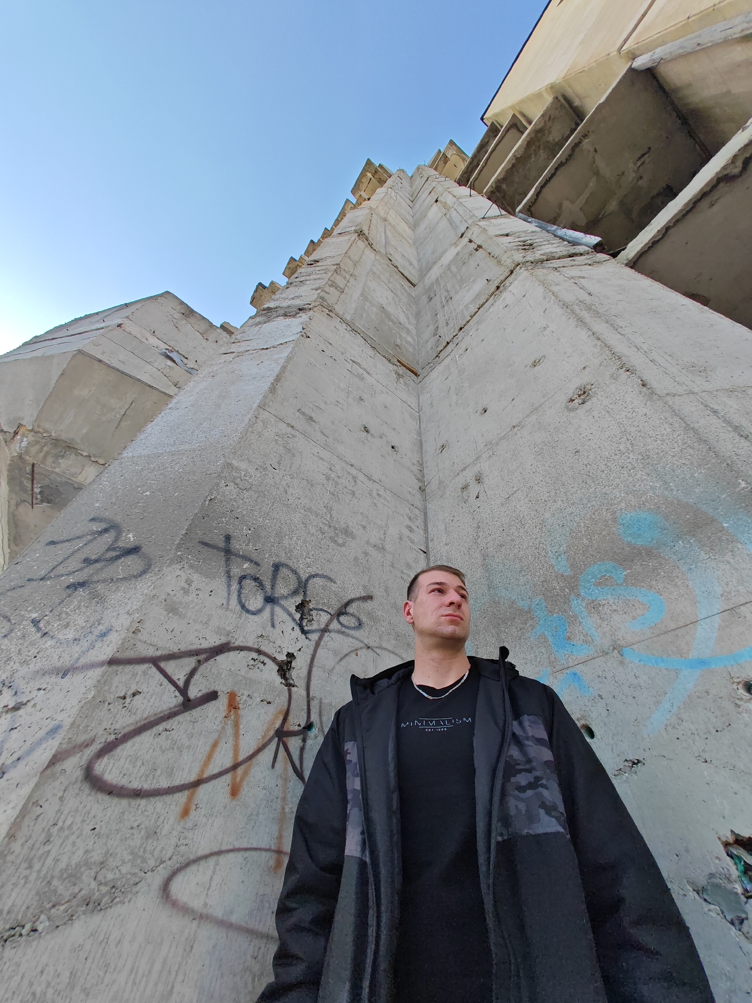 The image captures a male individual standing at the base of the Monument to 1300 Years of Bulgaria, located on the Shumen Plateau in Shumen, Bulgaria. The perspective is low-angle, looking upwards at the massive, angular, unpainted concrete structure, which displays characteristic Brutalist architecture. The concrete surfaces are weathered, with visible cracks, some exposed rebar, and extensive graffiti in black, blue, and brown tones.
The male subject, appearing to be in his twenties, has short, light brown hair. He is positioned in the lower-right section of the frame, facing slightly to his left and looking upwards. He wears a black long-sleeved shirt with the text "MINIMALISM EST. 1980" on the chest, beneath an open black hooded jacket featuring dark camouflage-patterned panels on the sides. A silver-colored chain necklace is visible around his neck. The sky in the upper portion of the image is clear and light blue. No other individuals or active interactions are visible.