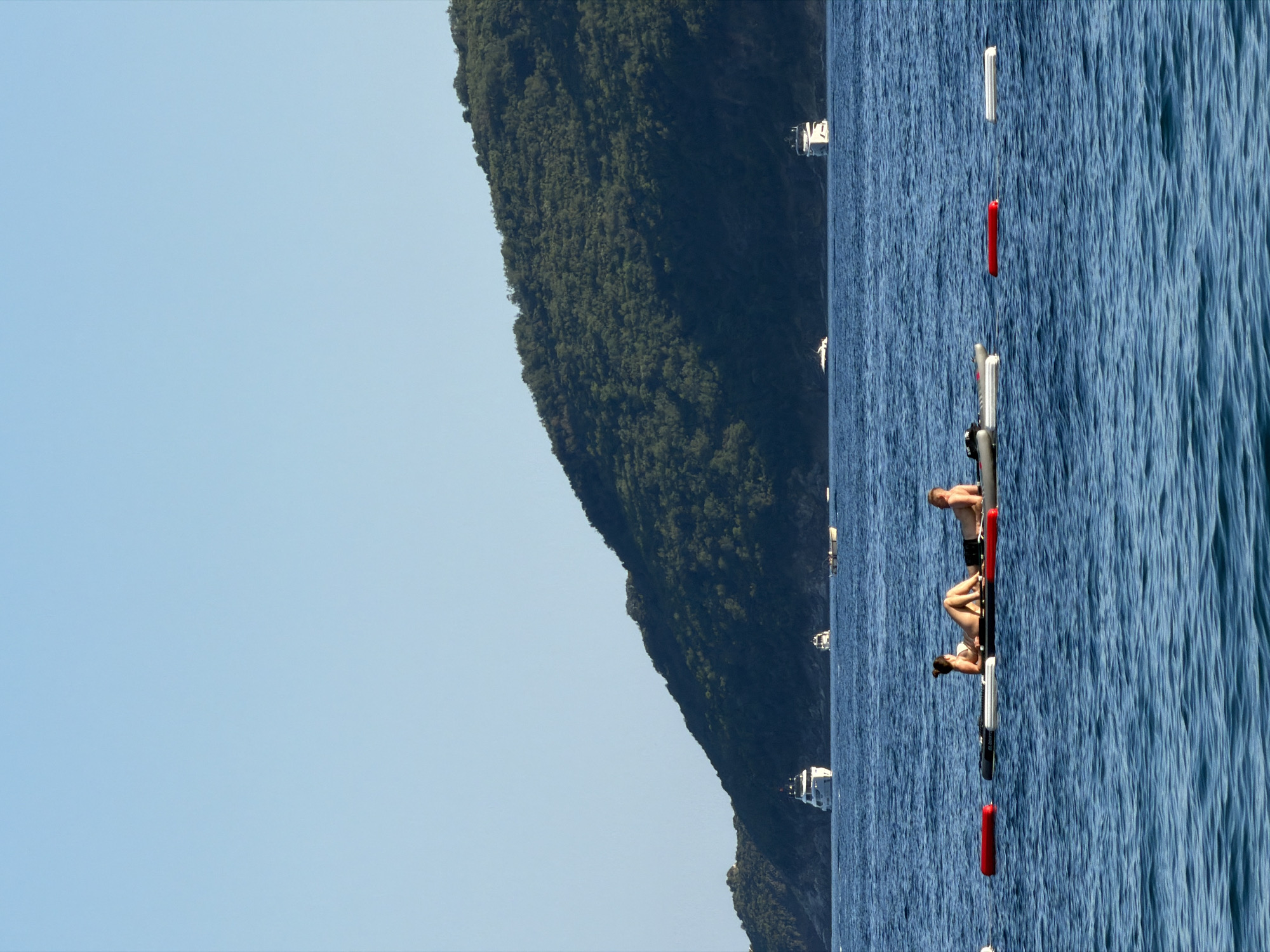 The image captures a sunny day at sea near Budva, Montenegro, with a backdrop of a lush green mountain slope. In the foreground, two people are seen relaxing on a floating mat, one lying on their back and the other on their side. They are positioned on the water with several buoys visible in the vicinity. Further out in the sea, several yachts are anchored, indicating a popular tourist destination. The sky is clear and blue, suggesting pleasant weather for outdoor activities.
