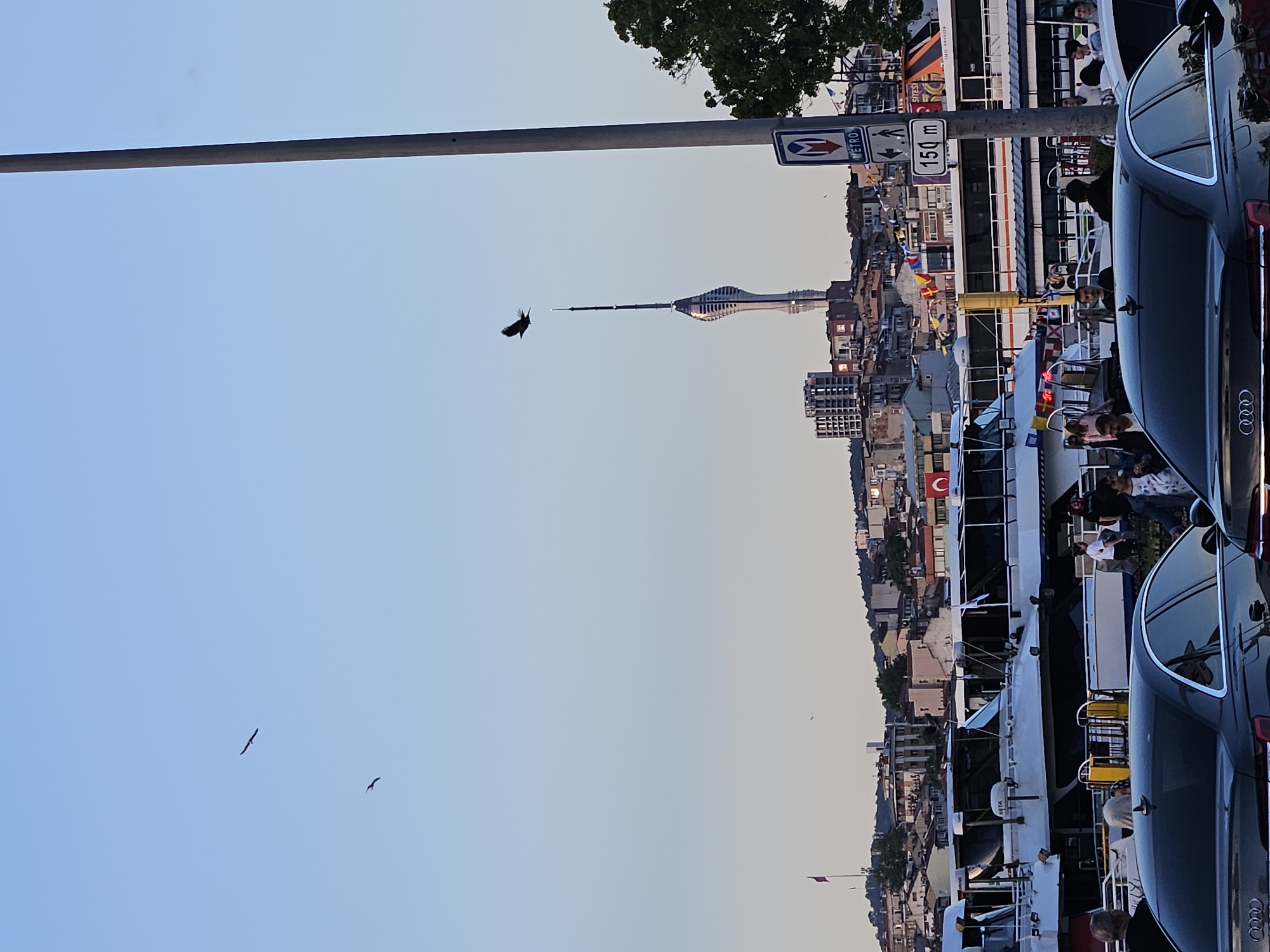 The image shows a sideways view of Kadıköy, Turkey, with the Istanbul TV Tower visible in the distance. A black Audi car is parked on the right side of the frame. The foreground features a ferry with passengers on board. A sign indicating "Metro" and a distance of "150 m" is present. Birds are flying in the sky. Buildings, houses, and the Turkish flag are visible in the urban landscape.