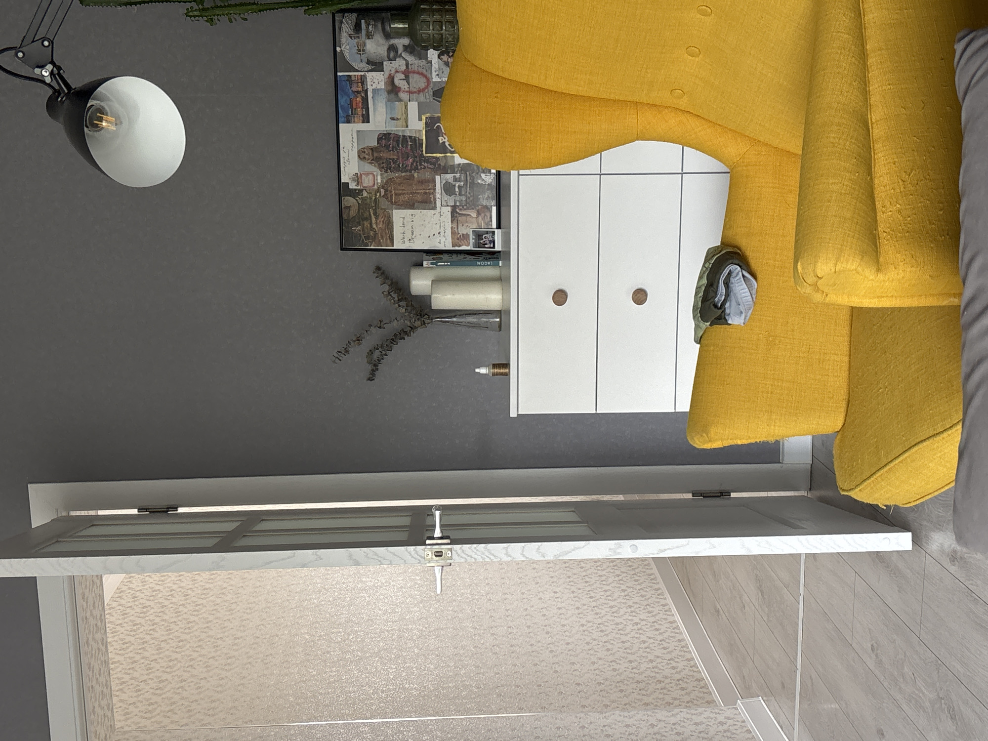 The image shows a room interior with a yellow armchair, a white cabinet, and a door leading to a room with light-colored flooring. A black adjustable lamp is positioned on the left wall, above a framed picture. The wall is painted gray. Some folded clothing rests on the side of the cabinet next to the armchair. There are no people visible in the frame. It is not possible to confirm the location in Chaikovsky, Russia, based on the given image.