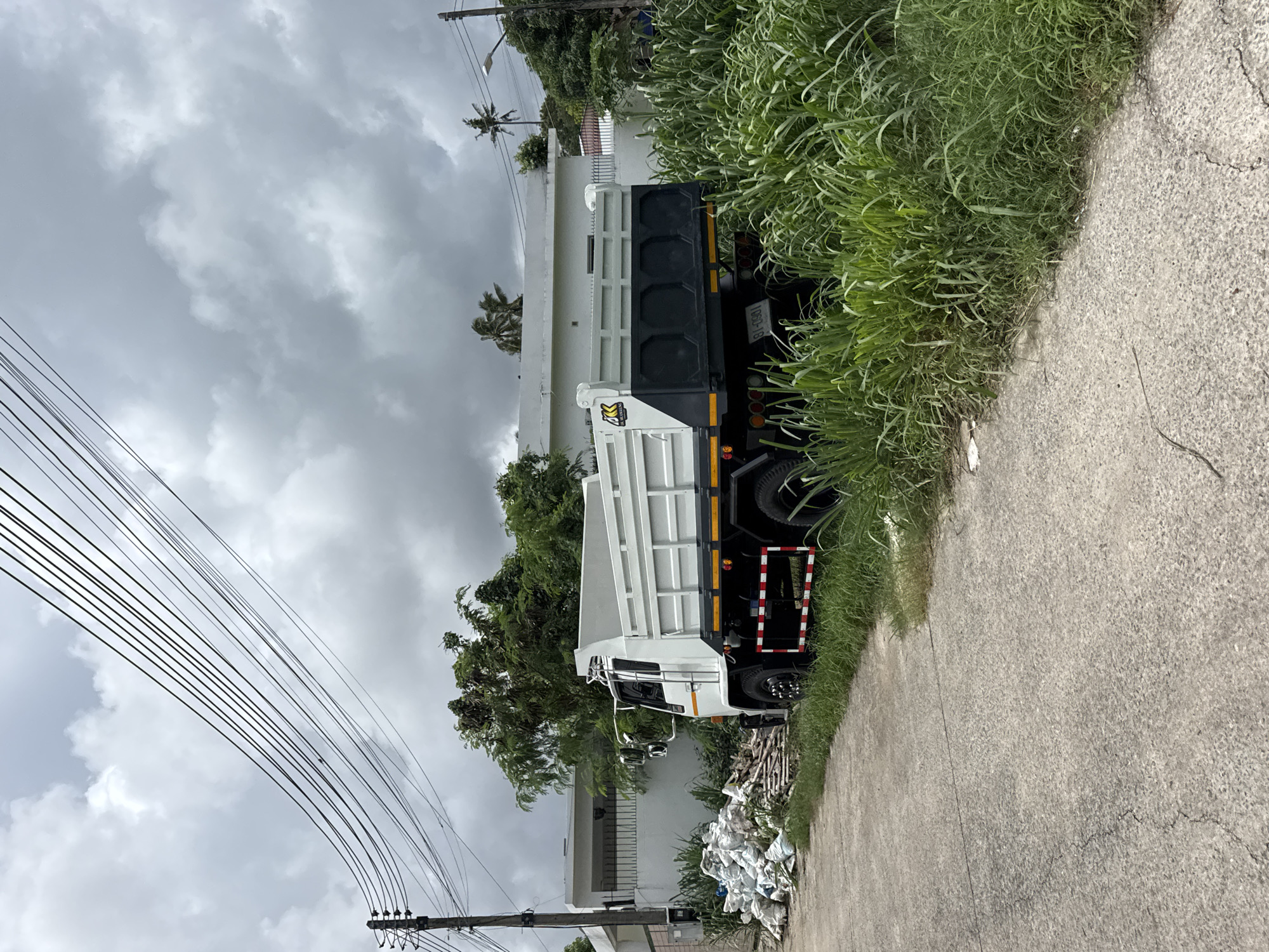 A white dump truck is parked on the side of a paved road, with its front facing left. The truck has a white bed with gray panels on the rear. A pile of white bags and some wooden planks are situated on the ground to the left of the truck, near a white building. Tall green grass is growing along the side of the road and obscures the lower portion of the truck. Overhead, electrical wires are strung across a cloudy sky. The scene appears to be in a semi-urban or rural area.