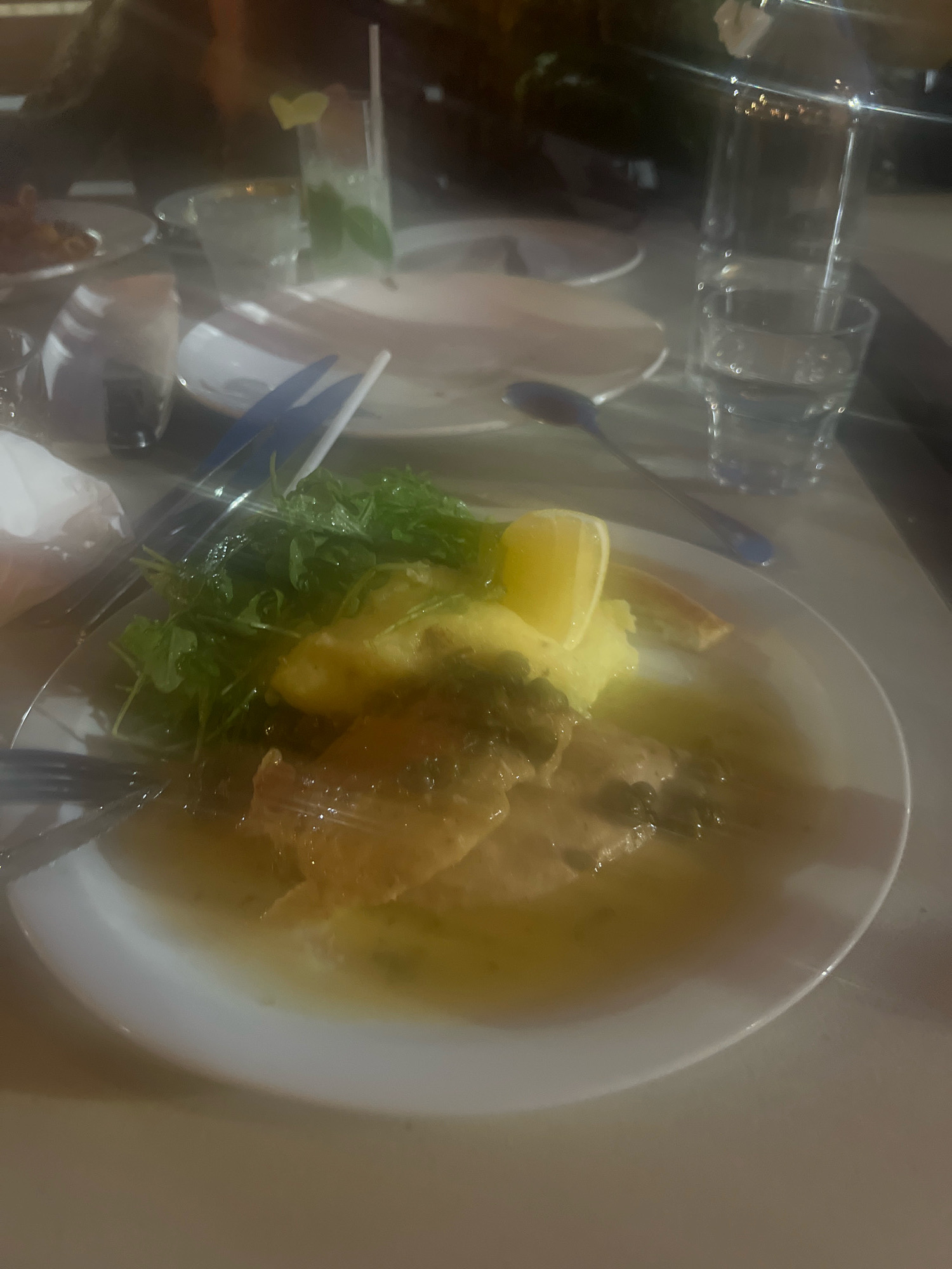 The image depicts a close-up, slightly blurry view of a meal served on a white plate. The meal appears to be fish or poultry in a light-colored sauce, accompanied by mashed potatoes and a green salad. A slice of lemon garnishes the dish. In the background, partially visible are drinks, possibly cocktails, and glassware, suggesting a dining establishment. The lighting suggests an indoor evening setting. There are no clear indicators of the specific location within Markham, Canada. The scene focuses solely on the presented food and accompanying beverages.