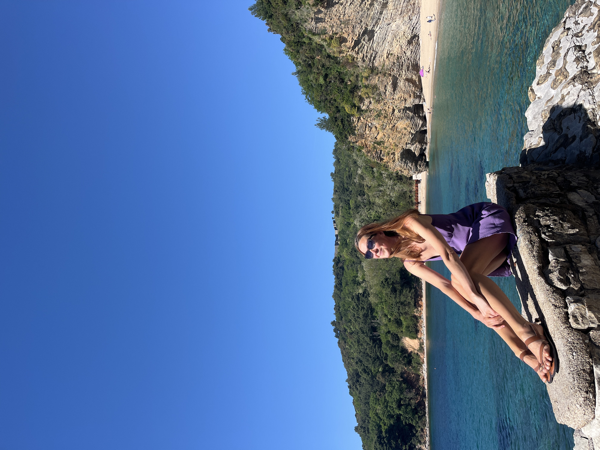 The image captures a woman sitting on a rocky outcrop overlooking a beach and clear turquoise water. The location appears to be near the city of Budva, Montenegro, given the coastal landscape, rocky cliffs, and vegetation characteristic of the region.
The woman is wearing a short purple dress and sunglasses. She's seated on the rocks, her legs bent and drawn up towards her. She wears brown sandals.
Behind her is a rugged cliff face covered in vegetation, dropping down to the beach below. The beach has a few people scattered along its length, and a couple of umbrellas provide shade. The water is clear and calm. The sky is bright and cloudless.