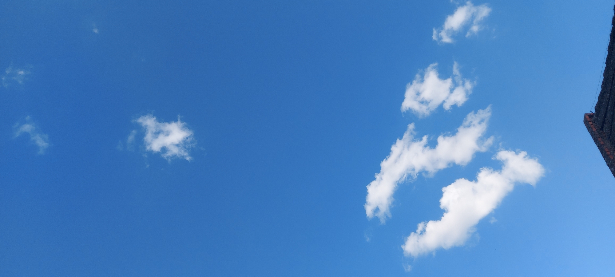 An image shows a clear blue sky with scattered white clouds. In the foreground, the corner of a dark-tiled roof is visible.