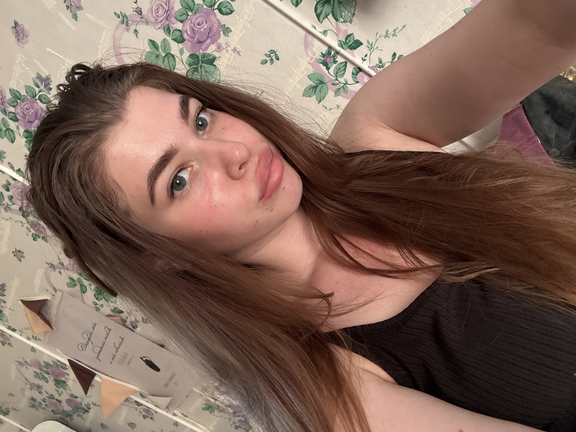 A young woman with long brown hair and blue eyes is posing for a selfie. She is wearing a black tank top. The background consists of a wall covered in a floral pattern with purple roses and green leaves. There is also a small decorative banner with text and small flags visible in the lower left corner. No information allows to locate this image in the city of Мещёры, Russia.