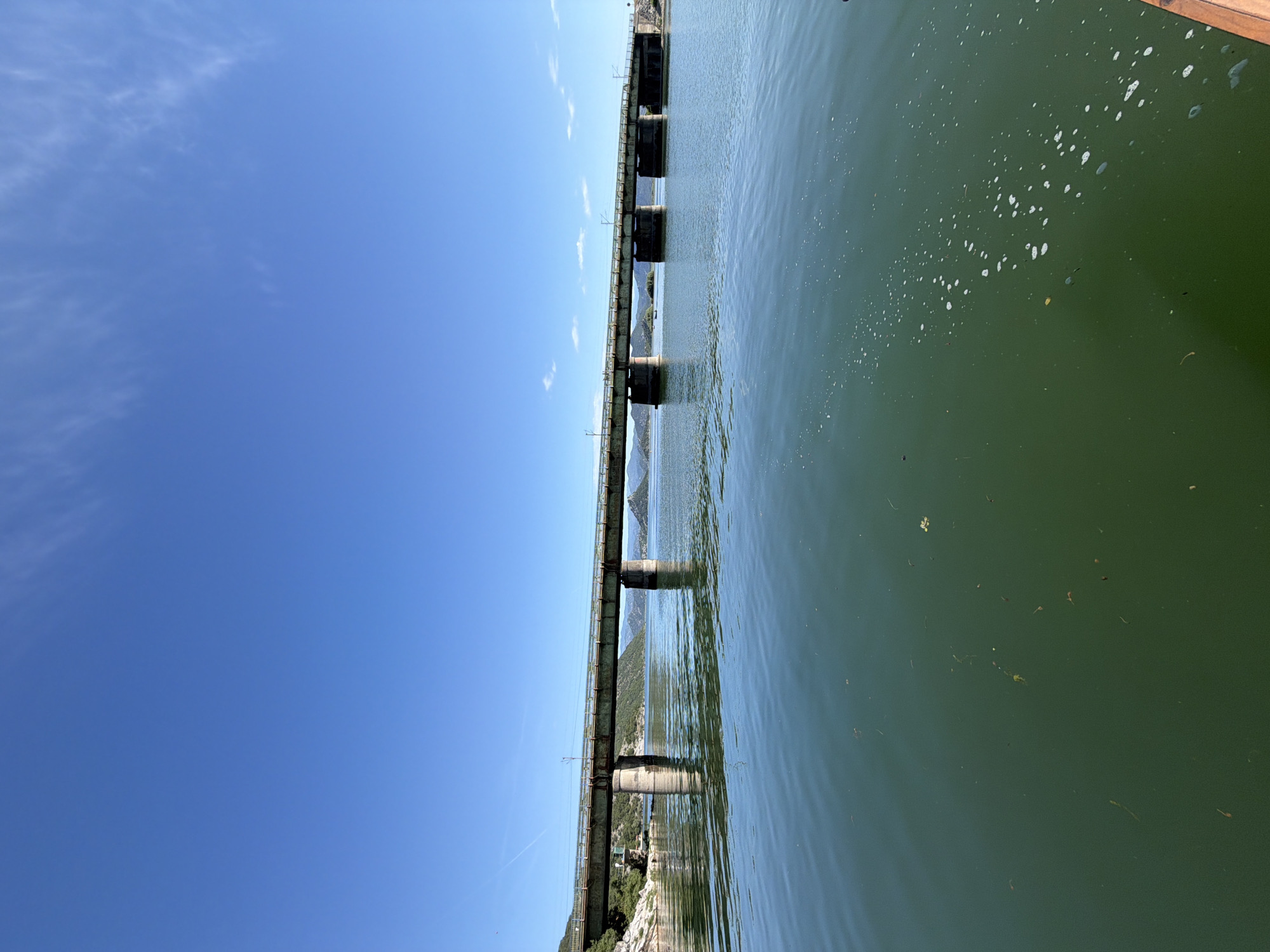 The image depicts a concrete and metal multi-span railway bridge traversing a wide body of calm, green-tinted water, under a clear blue sky with minimal cirrus clouds. The scene is located in Kruševica, Montenegro. The bridge, which appears weathered with a greenish-grey hue, is supported by several robust piers visible in the water, creating reflections below. Overhead lines, characteristic of electrified railways, are discernible along the bridge's structure. To the left, a vegetated, rocky shoreline meets the water, extending into distant mountains that frame the horizon. The water surface in the foreground contains scattered organic debris and patches of white foam or bubbles, particularly on the right side. A small segment of a wooden structure, possibly a boat or dock, is partially visible in the bottom right corner. No people or active vehicles are present in the frame.