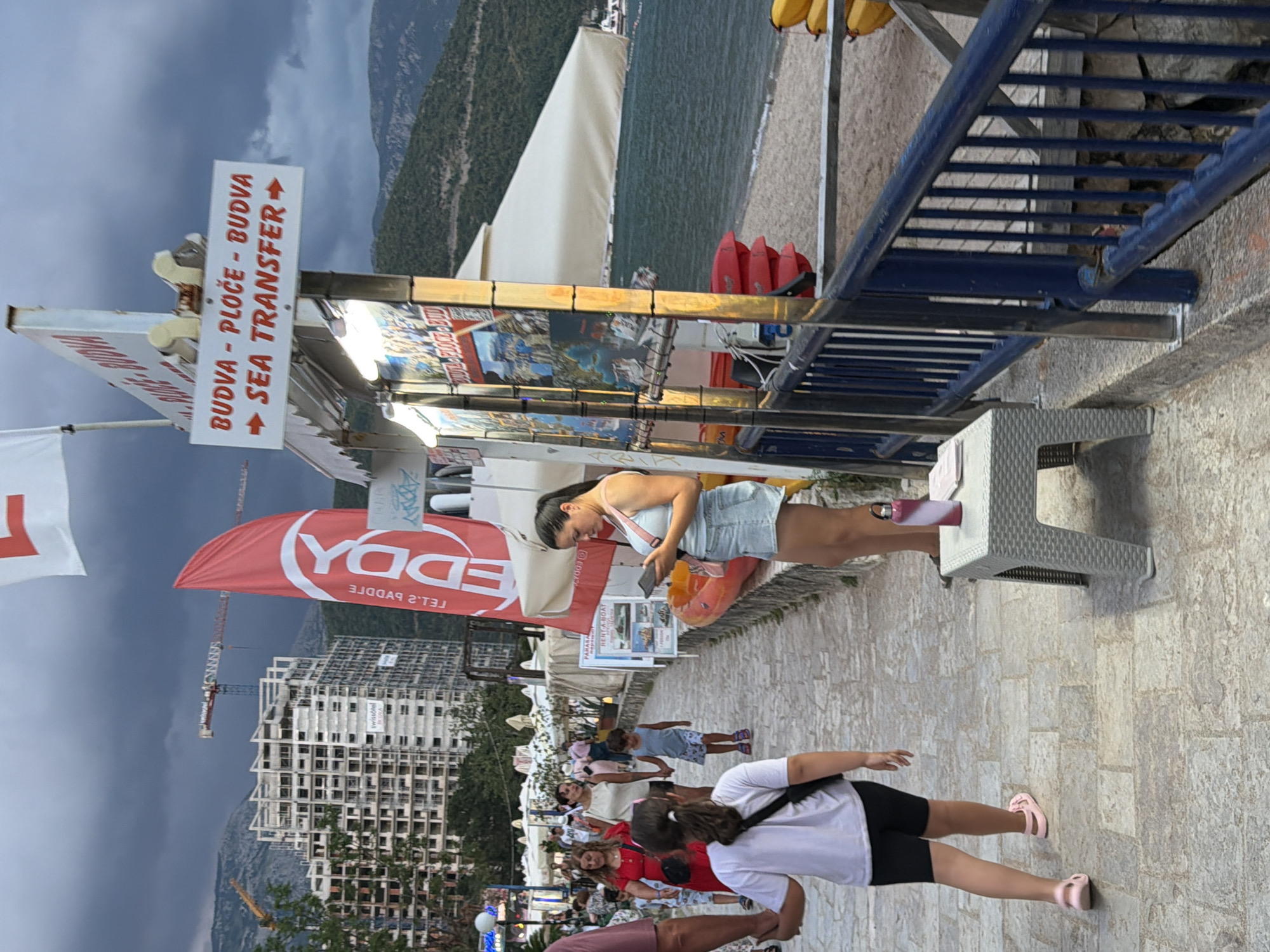 A woman stands on a paved walkway next to a beach in Budva, Montenegro, looking at her phone. She is wearing a light blue tank top and denim shorts. To her left is a small table with a water bottle and some papers. Behind her is a sign advertising "SEA TRANSFER" to Budva and Ploče. Further back, people are walking on the path, and there are buildings under construction and a crane in the background. The sea is visible to the right, with a beach and kayaks. The sky is cloudy.