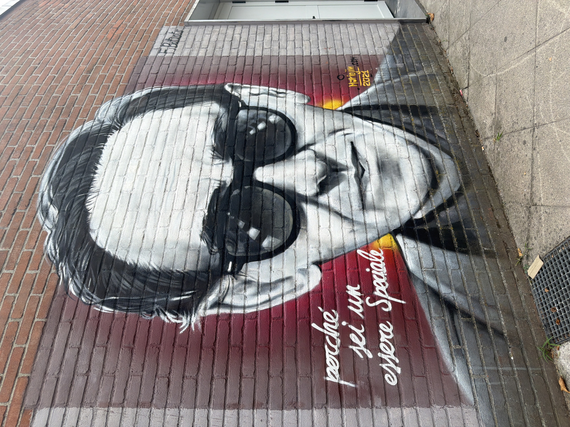 The image shows a mural painted on a brick wall in Corsico, Italy. The mural depicts a black and white portrait of a man wearing large sunglasses. To the left of the portrait, in white spray paint, are the Italian words "perché sei un essere Speciale", which translates to "because you are a special being". In the lower right corner of the mural, there is a signature that reads "Marijini.com 2021". The wall is made of red brick, with some sections painted grey and red. A white door is visible to the right of the mural. The ground in front of the wall is paved with concrete tiles and a metal grate is present.