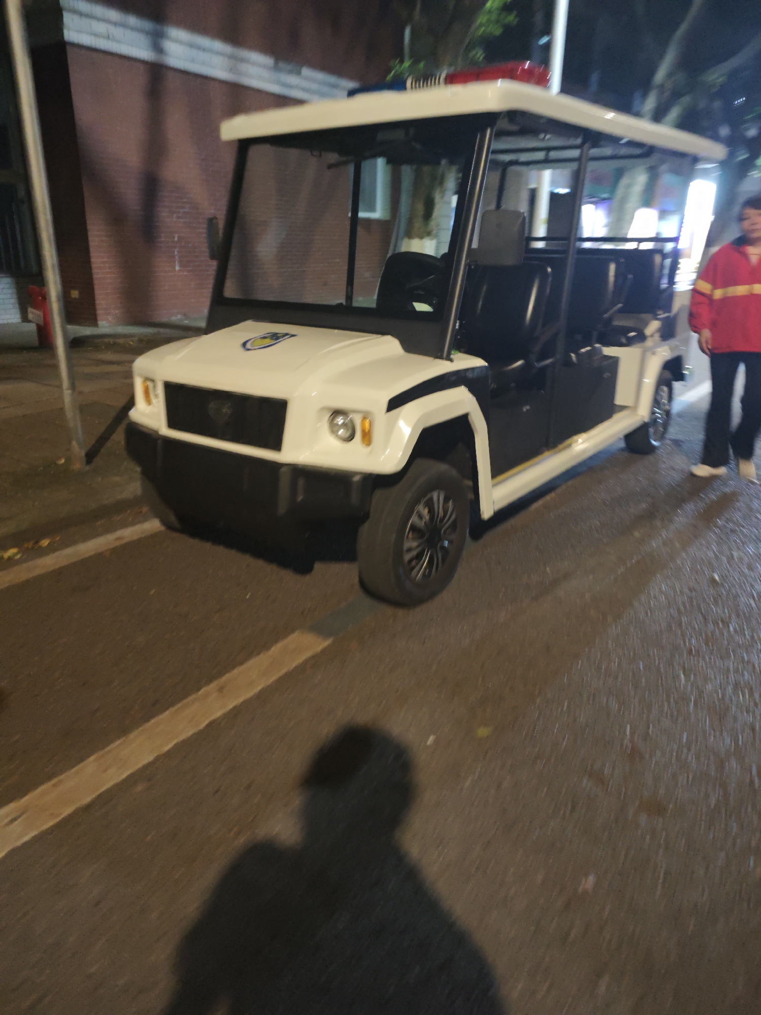 The image depicts a white and dark gray golf cart-like vehicle with a red and blue emergency light bar on the roof. The vehicle is parked on a paved surface with a yellow line. A person in a red jacket and dark pants is standing near the rear of the vehicle. The surrounding environment includes a building with a brick facade and street lighting. The photo was taken at night in Shapingba District, China.
