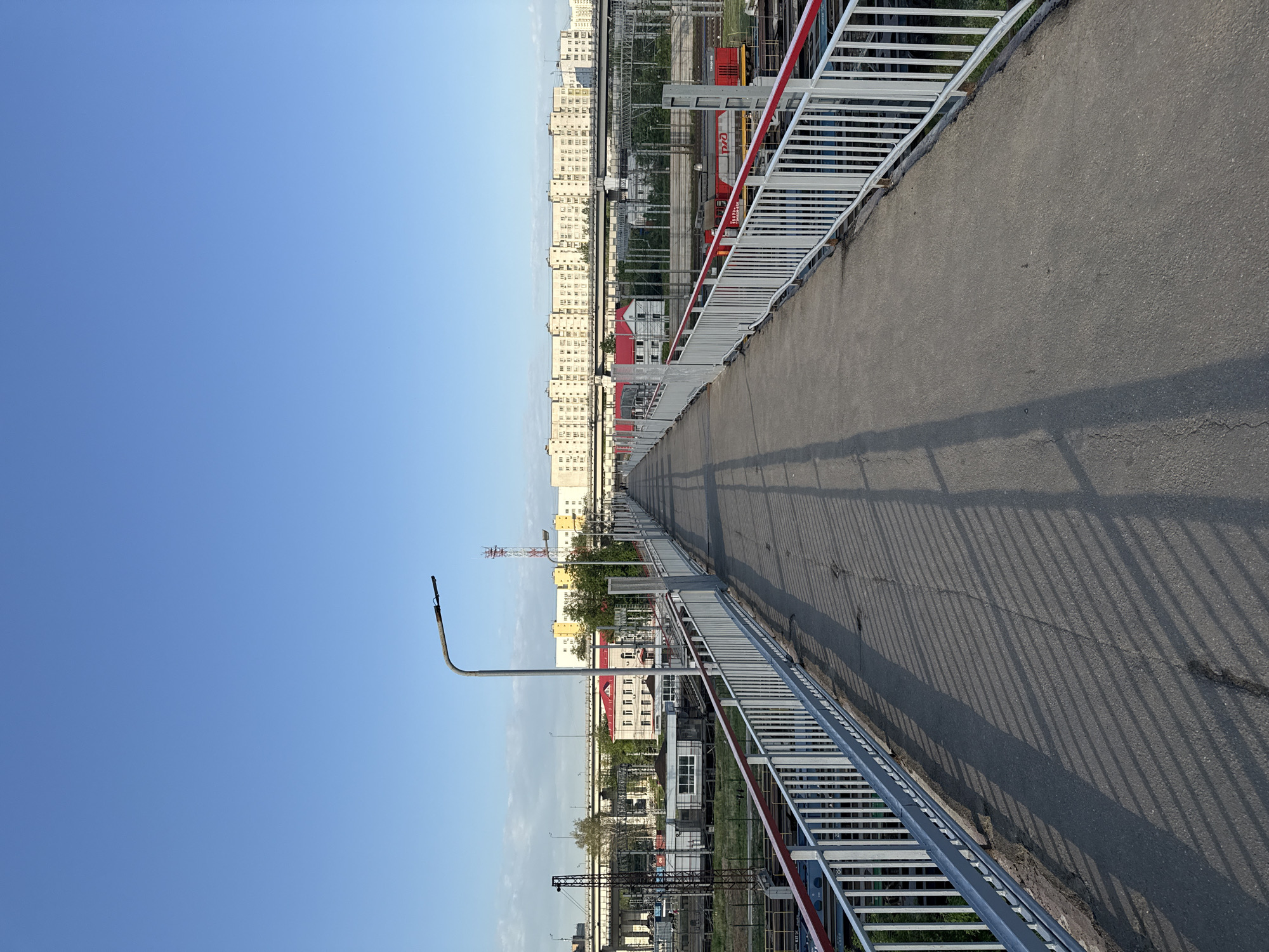 The image depicts an elevated pedestrian walkway in Moscow, Russia. The walkway has a grey asphalt surface and is bordered by white metal railings with red accents. Shadows from the railings are cast onto the walkway surface. To the left of the walkway, the sky is a clear, bright blue. On the right side, a cityscape is visible, featuring multi-story apartment buildings with light-colored facades. Closer to the walkway are industrial structures, railway tracks, and vegetation. A street light is visible in the lower-left. A red train is visible with the 'PЖД' logo.