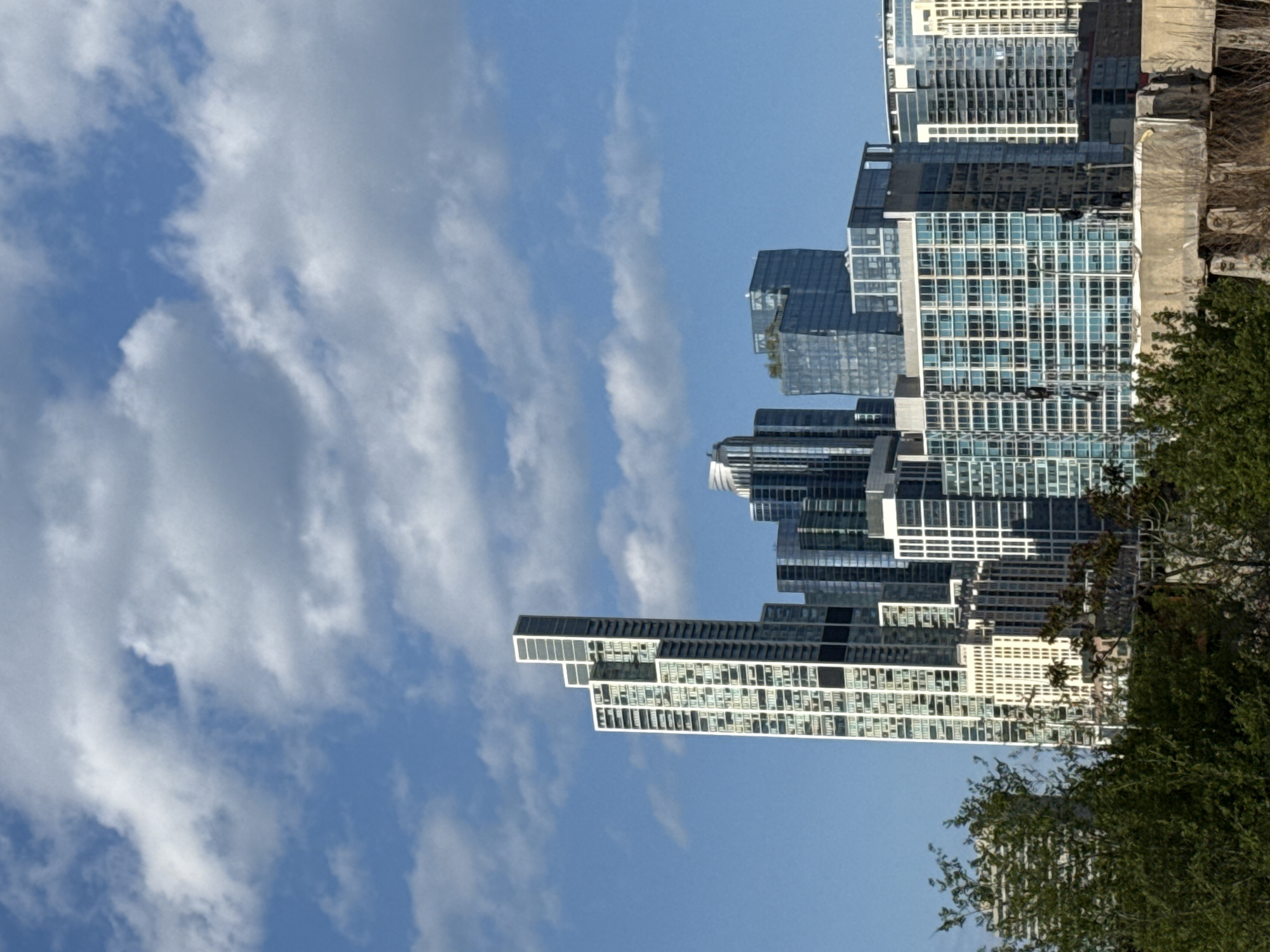 The image captures a view of the Chicago skyline against a partly cloudy sky. The buildings are modern, with glass and steel facades, and are densely packed together. The clouds are scattered and white, contrasting with the blue sky. Some greenery, likely trees, is visible on the right side of the frame. The image shows an upward angle, emphasizing the height of the buildings.
