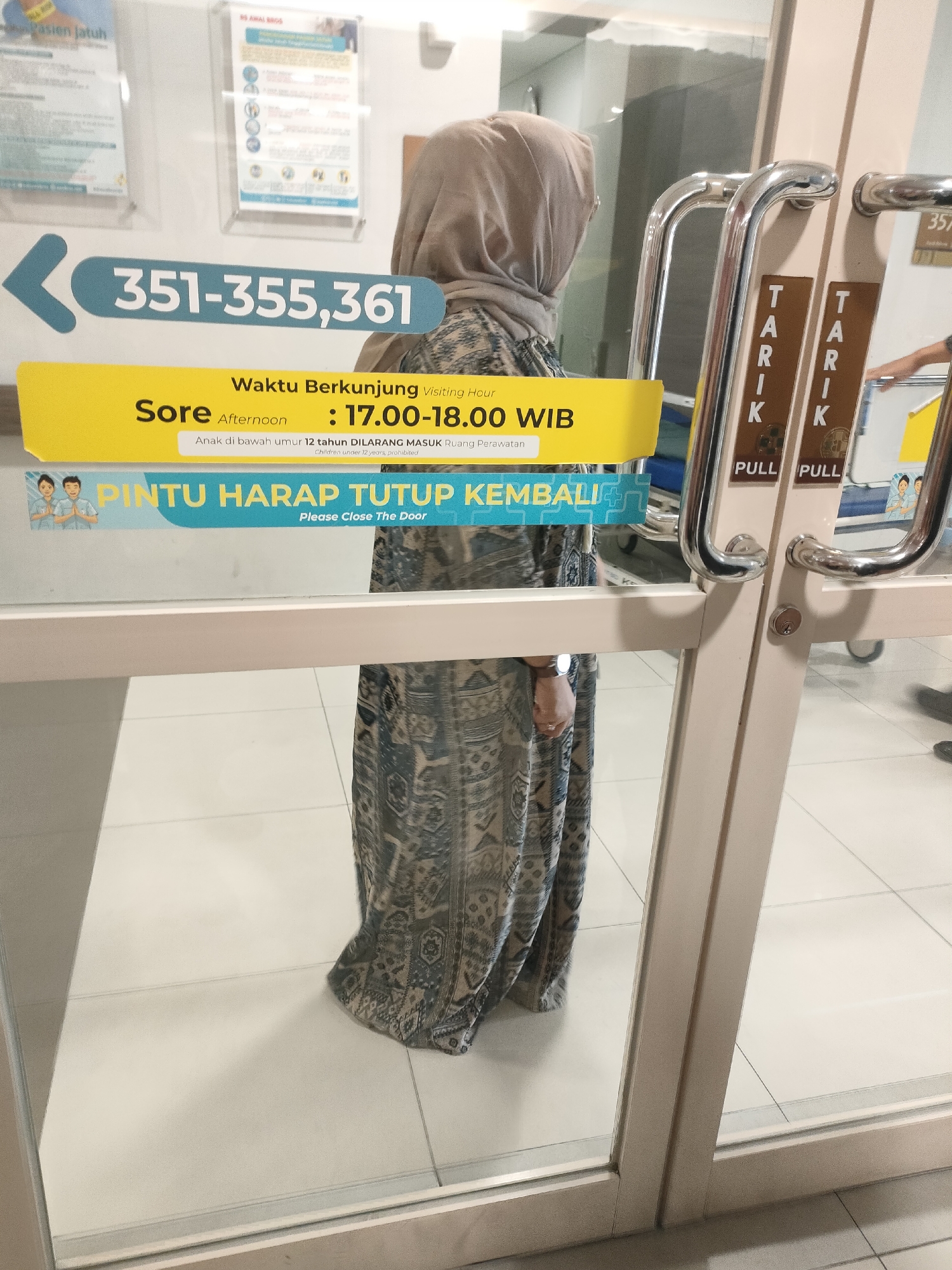 The image shows a hospital hallway with glass doors. A woman wearing a hijab and a patterned dress stands in front of the doors, her reflection visible in the glass. Signs on the doors indicate visiting hours and instructions to close the door. The hallway floor is tiled, and a sign with a phone number is visible on the left side of the image. The hospital appears to be a modern facility.
