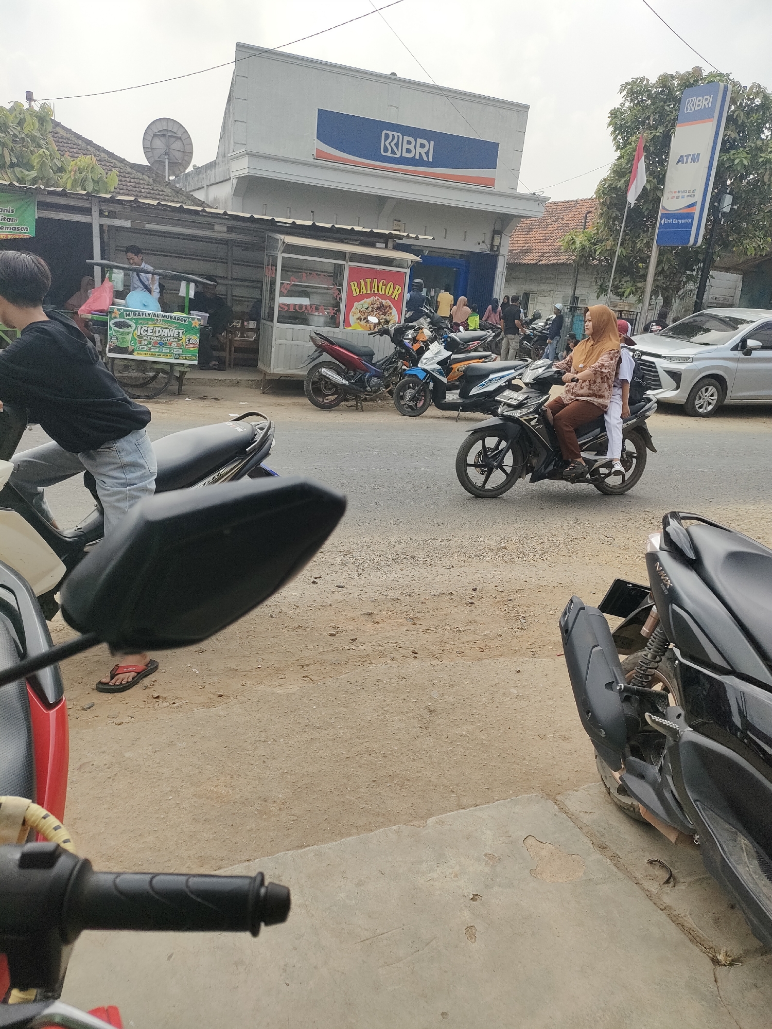 In Sukoyoso, Indonesia, a scene unfolds along a street lined with low-rise buildings and trees. A prominent BRI bank with an ATM sign stands out. Various motorcycles are parked and in motion, indicating a bustling area. A woman wearing a hijab and a young person in a white uniform ride a black motorcycle. In the foreground, the handlebar and side mirror of a motorcycle are visible. To the left, a food stall with a sign advertising "ICE DAWE" and "BALAGOR" is set up, with people around it. A silver car is parked to the right of the scene. The overall environment suggests a typical street scene in a populated area of Indonesia.