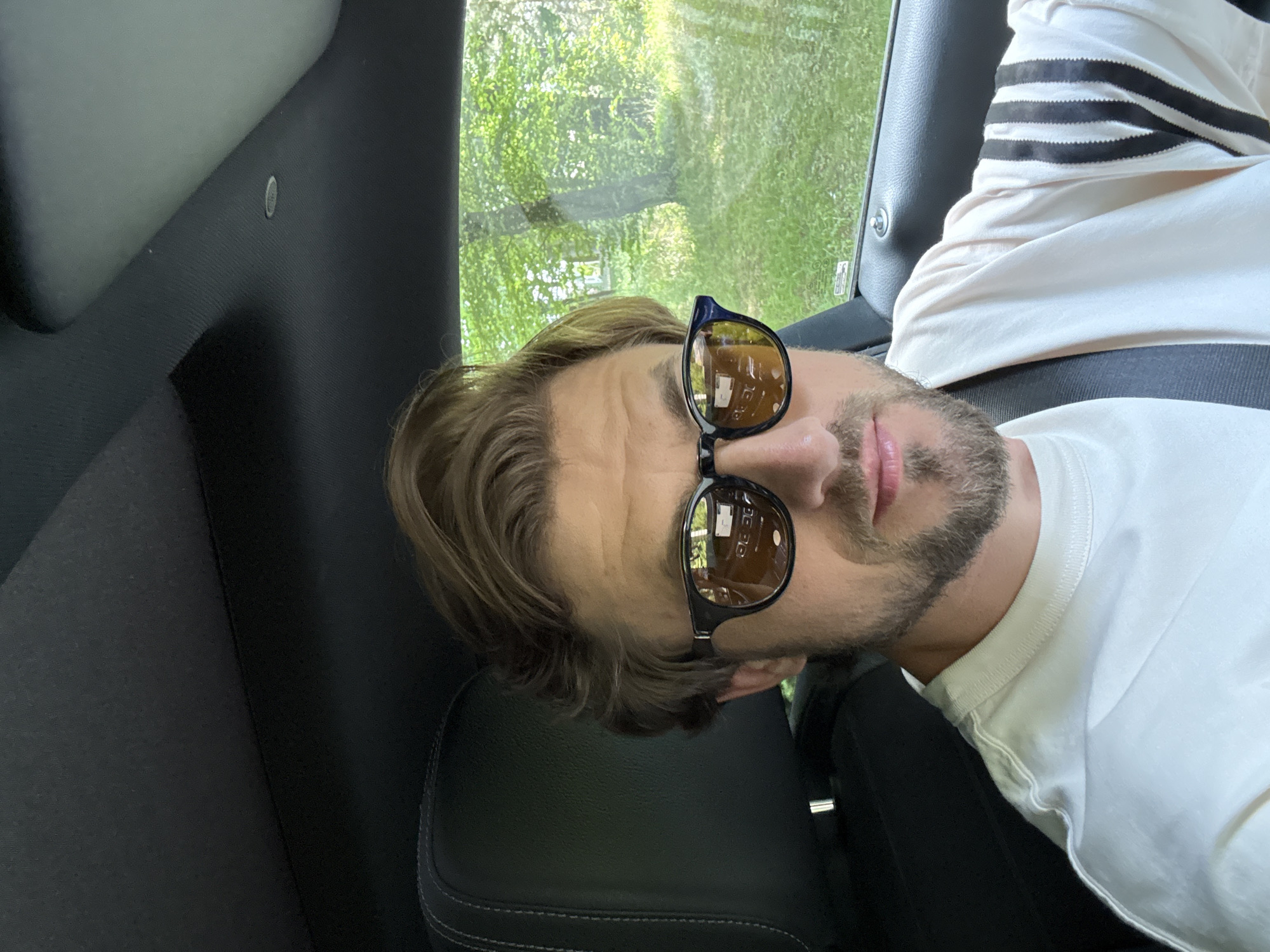 The image shows a close-up, portrait-style shot of a man wearing sunglasses and a white t-shirt with black stripes on the sleeves. He is inside a vehicle, with the interior upholstery and a window with greenery visible in the background. The man has light brown hair and a beard. He appears to be looking towards the camera, with his head tilted. The scene suggests a casual, possibly daytime setting, given the natural light and the greenery outside. The image is oriented sideways. There is no visible text in the image. The location context provided is Krubki-Górki, Poland.