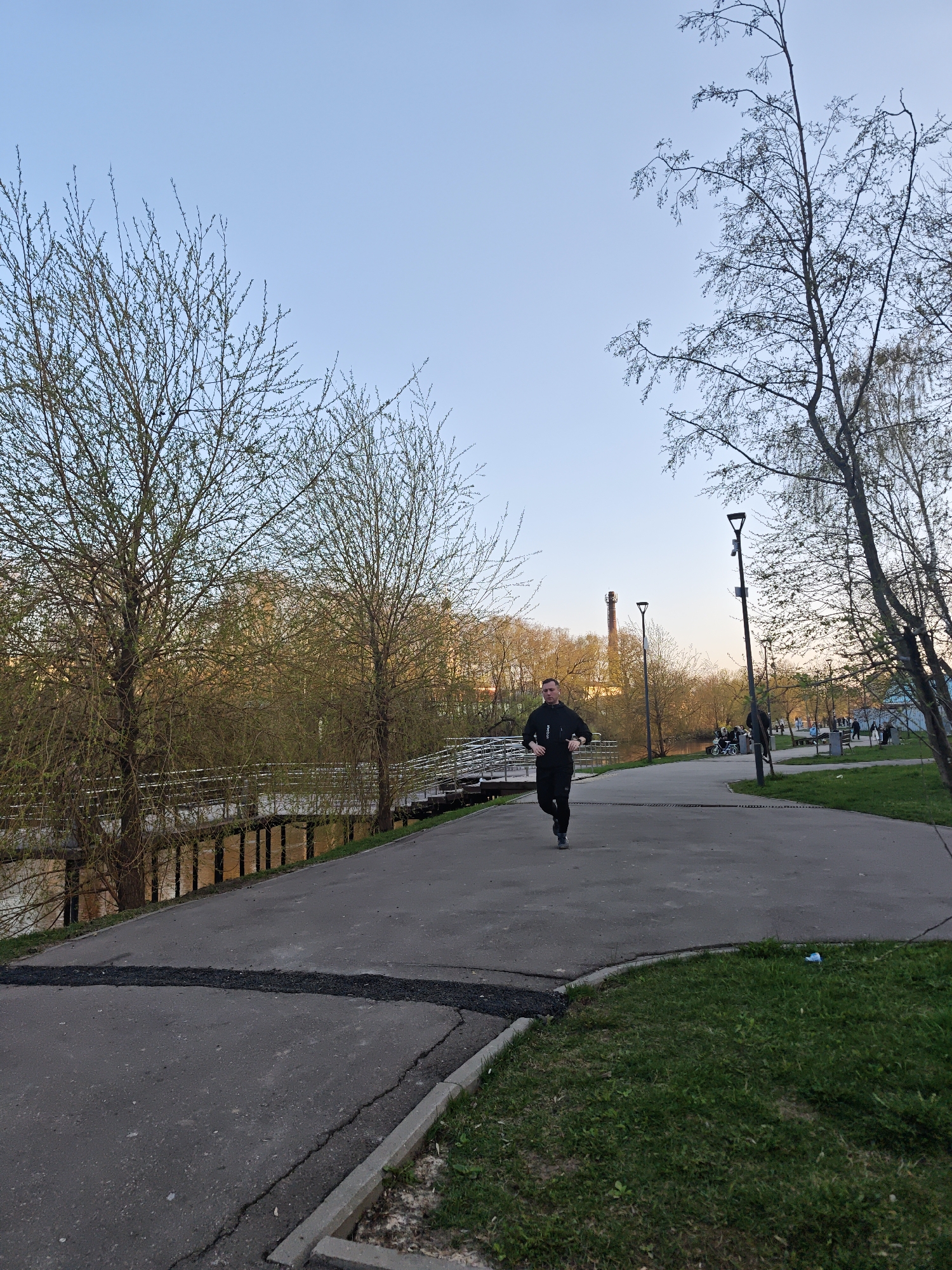 A man is jogging on an asphalt pathway in a park. The pathway is bordered by a river on one side, with a metal railing along the edge of the path beside the river. On the other side of the path is a grassy area. In the background, there are trees, lampposts, and people strolling. A tall chimney structure is visible in the distance. The location is the city of Noginsk, Russia. The time of day appears to be late afternoon or early evening, based on the lighting.