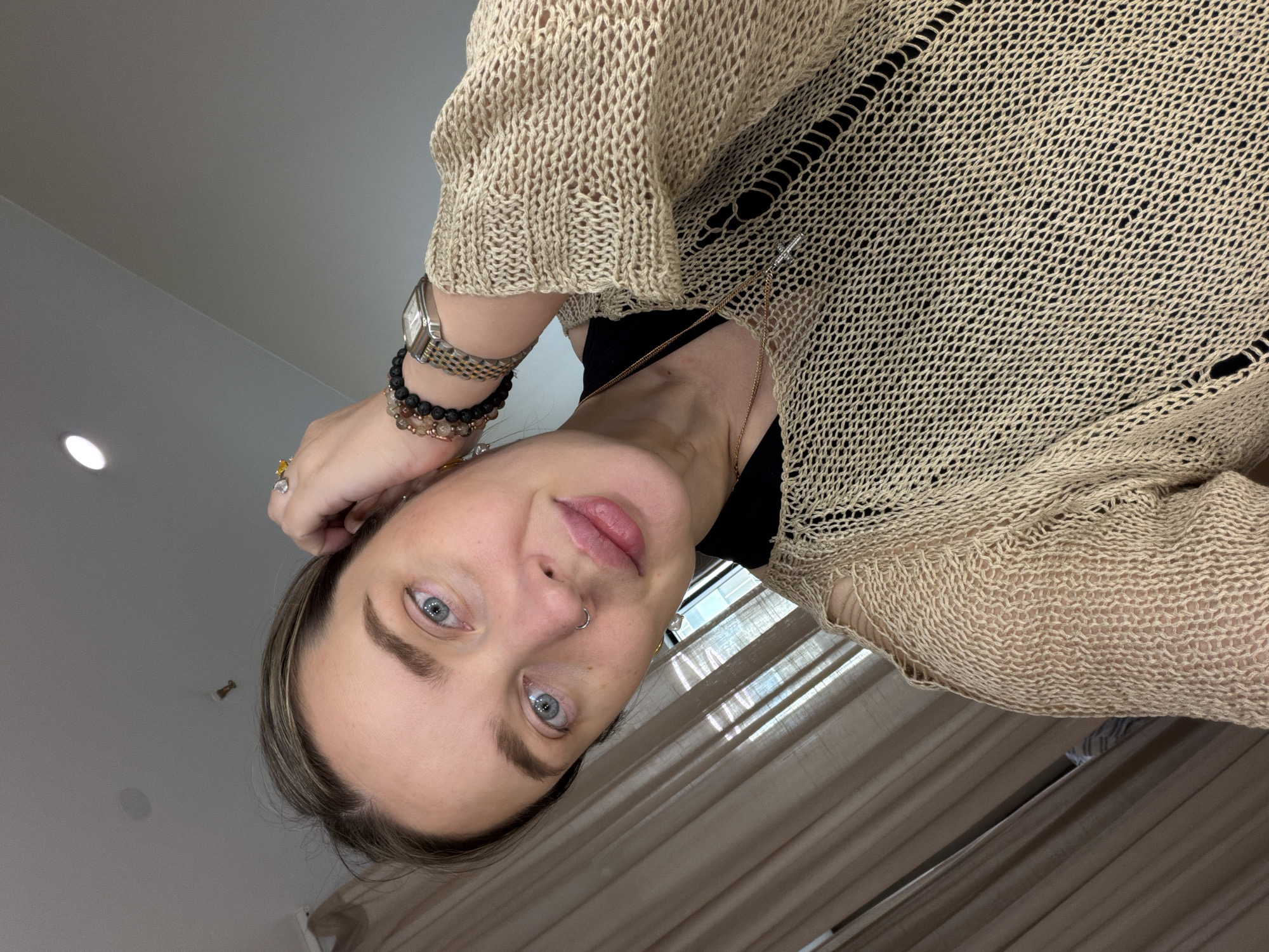 A woman with blue eyes and fair skin is seen in a close-up, tilted shot. She is wearing a beige, loosely-knit sweater over a black top. Accessories include a silver wristwatch, multiple beaded bracelets, a gold necklace with a small cross pendant, and a ring on her finger. A small hoop earring is visible in her nose. The background includes a white ceiling with a recessed light fixture, as well as beige curtains. The location is presumed to be indoors, but specific city and location details are unknown.