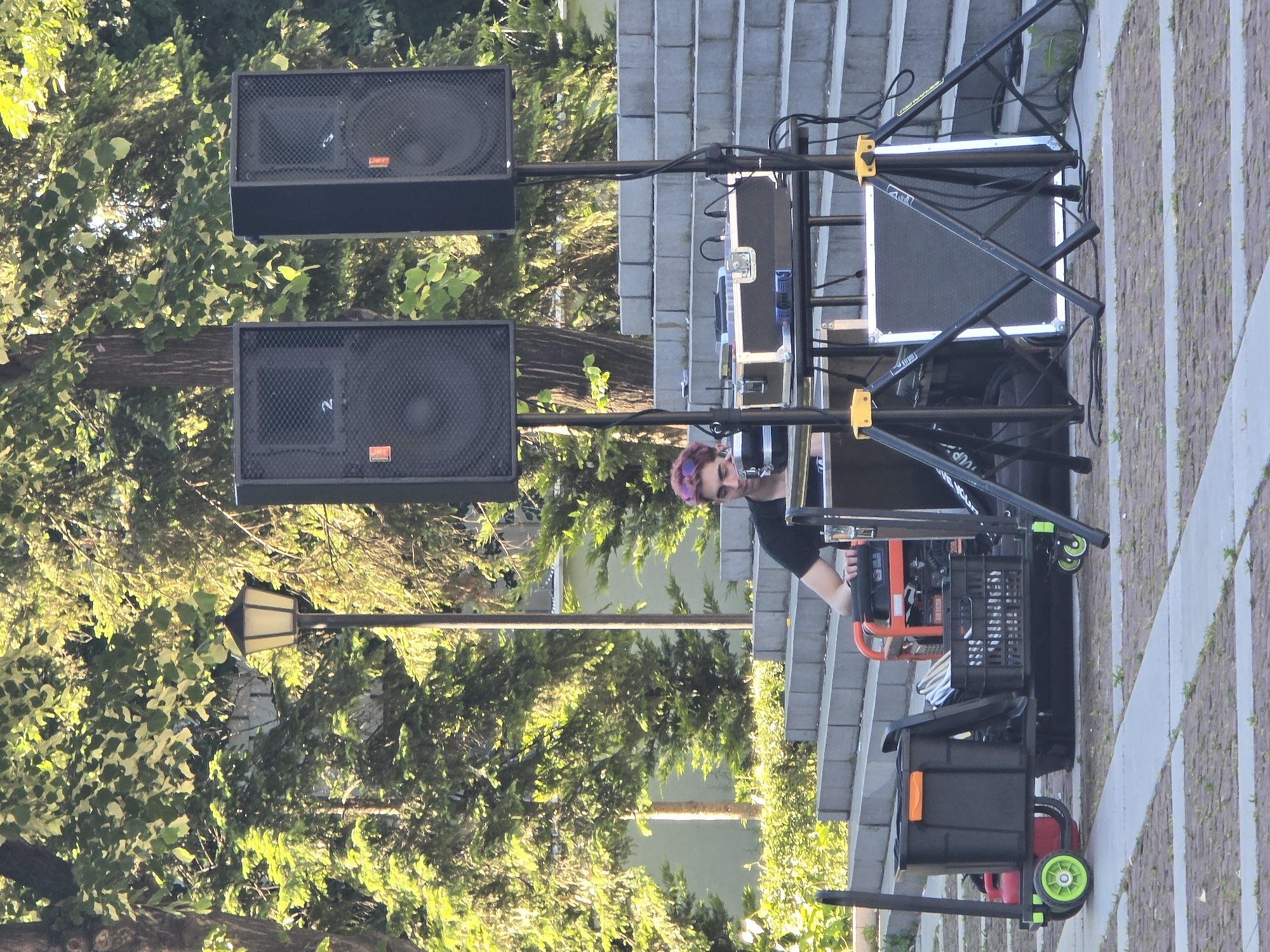 A person with pink hair stands behind a black metal scaffolding with two large black speakers elevated on poles to the left. A portable generator is located to the right, next to a black rolling utility cart. The scaffolding is positioned on a set of grey stairs. In the background, trees with green leaves are visible. In the distance are dark street lights and a building. The person appears to be setting up audio equipment outdoors in an urban setting. The pavers on the ground are patterned.