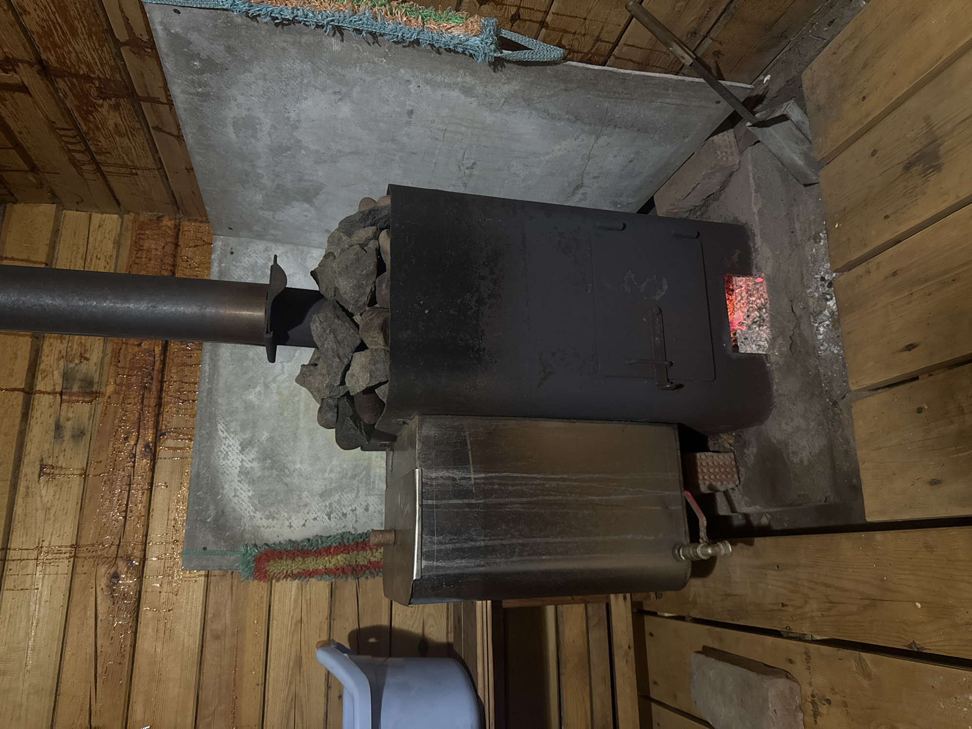 The image depicts a sauna stove in a wooden structure, likely a sauna room. The stove is black and contains rocks on top, presumably for heat retention. The firebox is partially visible, showing glowing embers inside. A metal pipe extends from the stove, likely for ventilation. Below the stove is a water heater, also made of metal. A plastic bucket sits on the floor near the heater. The wooden walls of the sauna are visible on either side, and a concrete or stone wall is behind the stove. Based on the prompt, the location is likely in the city of Mayak, Russia.