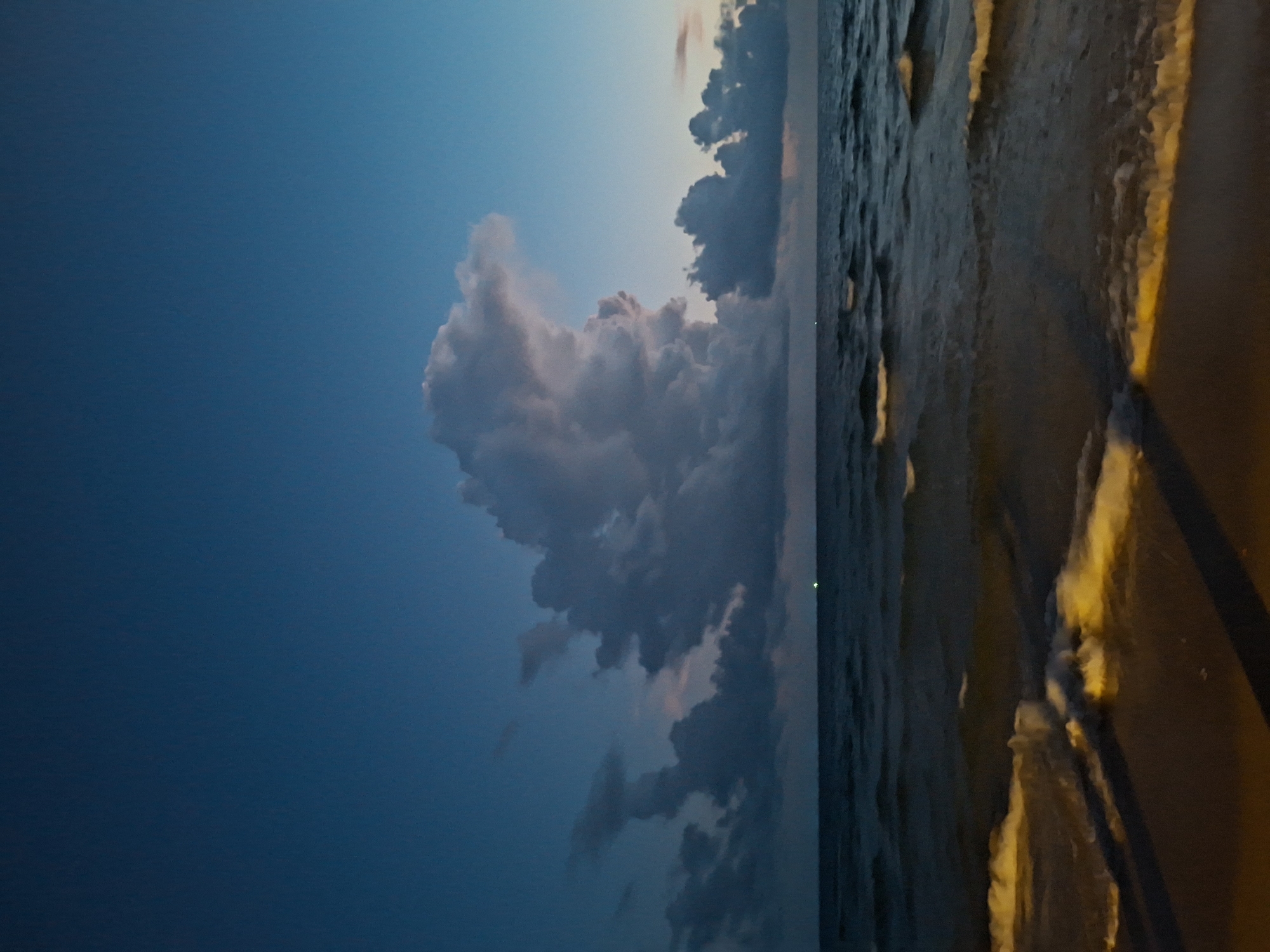 This is a vertical, low-angle, full shot of the ocean at dusk. The sky is a deep, gradient blue, with lighter shades near the horizon. A large, dramatic cumulus cloud formation is centered in the upper half of the frame, with softer, darker clouds to either side. The ocean is dark and choppy, with small waves breaking on a sandy shore in the foreground. The waves have a yellowish glow from artificial lighting, casting long shadows across the sand. There is a single, small green light visible on the horizon in the middle distance. No people or structures are visible.