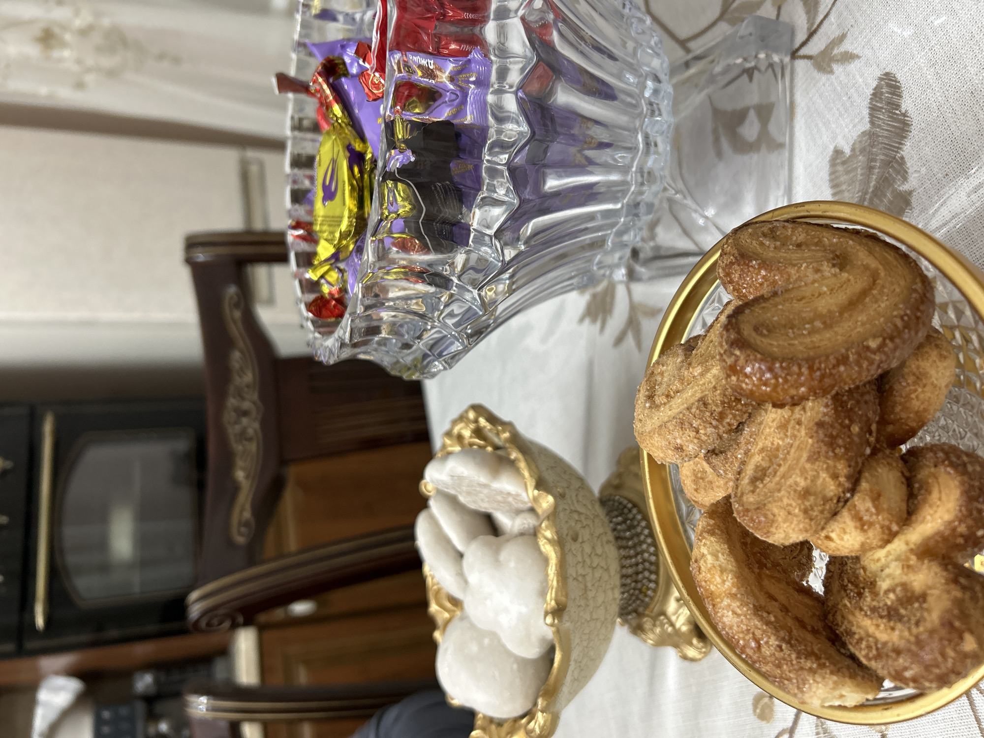 In the city of Nizhnee Kazanishche, Russia, a table is set with three bowls of sweets. In the foreground, a golden-rimmed bowl holds palmiers, a type of puff pastry cookie dusted with sugar. To the left, a decorative, ornate bowl displays white, soft sweets, possibly cookies or candies. A large, crystal glass bowl sits to the right, filled with individually wrapped chocolates in various colors, including purple, yellow, and red. The table is covered with a white tablecloth featuring a subtle floral pattern. In the background, a dark wooden dining chair is partially visible, and behind it, a black oven is mounted above kitchen cabinetry. The lighting suggests an indoor setting, likely a home dining area.