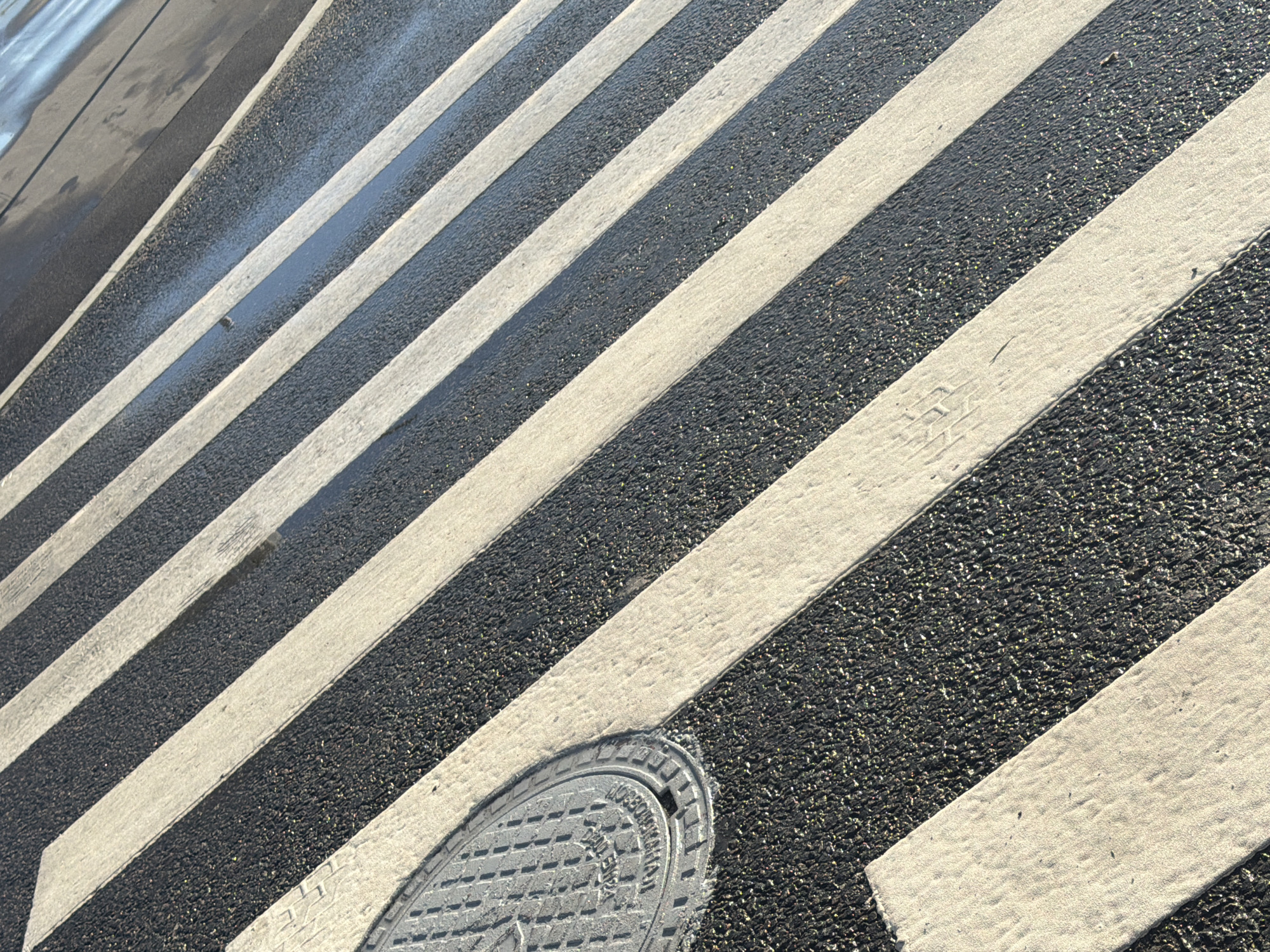 The image displays a section of a wet crosswalk, characterized by alternating black asphalt and white painted stripes. The stripes are angled diagonally across the frame, creating a sense of depth and perspective. The asphalt surface appears dark and reflective due to the presence of water, with small, shimmering particles embedded within it. The white stripes show some wear and tear, with subtle texture and faint tire marks visible on some.
In the bottom right corner of the frame, a circular manhole cover is partially visible. It is made of metal and has a textured surface with raised patterns and some illegible text inscribed on it. The overall scene suggests an outdoor urban environment, likely a street or road, possibly after rainfall. The lighting indicates it is daytime, with the sun casting reflections on the wet surface. No people are present in the frame. The mood is neutral, focusing on the texture and patterns of the road surface. The primary subjects are the crosswalk markings and the manhole cover, set within the context of a wet, paved ground.