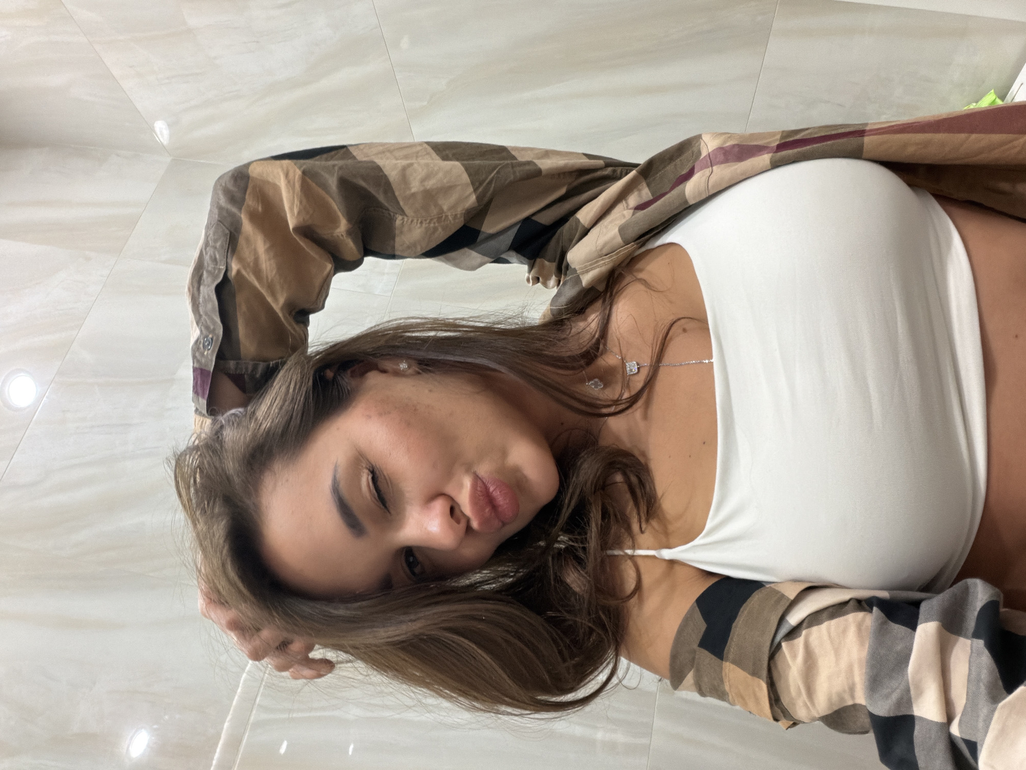 A woman is lying on her back in what appears to be a bathroom with light-colored tiled walls. She is wearing a white top and a plaid shirt. She has brown hair and is winking and pouting her lips towards the camera. A silver necklace is visible on her chest. No location is discernible from the image.