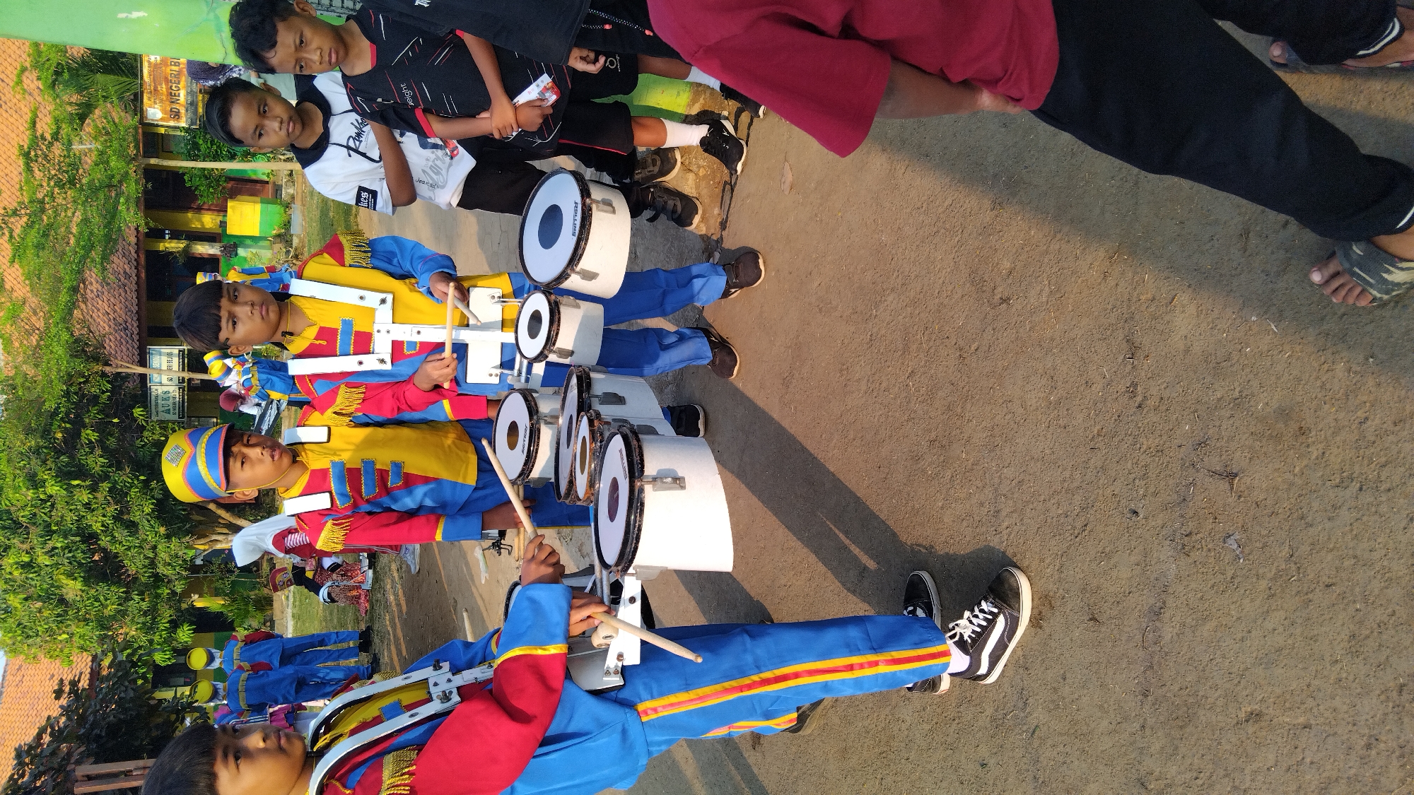 In Sembojo, Indonesia, on a dirt ground with a building in the background, a group of children are participating in a drum corps activity. Two boys in the foreground are dressed in colorful marching band uniforms, featuring red, yellow, and blue jackets with gold trim, and matching blue pants with yellow stripes. They are holding drumsticks and playing white snare drums attached to their chests with straps.

Behind them, several other children are visible. Some are also in similar marching band uniforms, while others are in casual clothing. One boy wears a white t-shirt with a graphic print, and another wears a black t-shirt with red accents. The background shows a building with a red-tiled roof and trees, suggesting an outdoor schoolyard or community space. The overall scene captures a moment of musical practice or performance by young students.