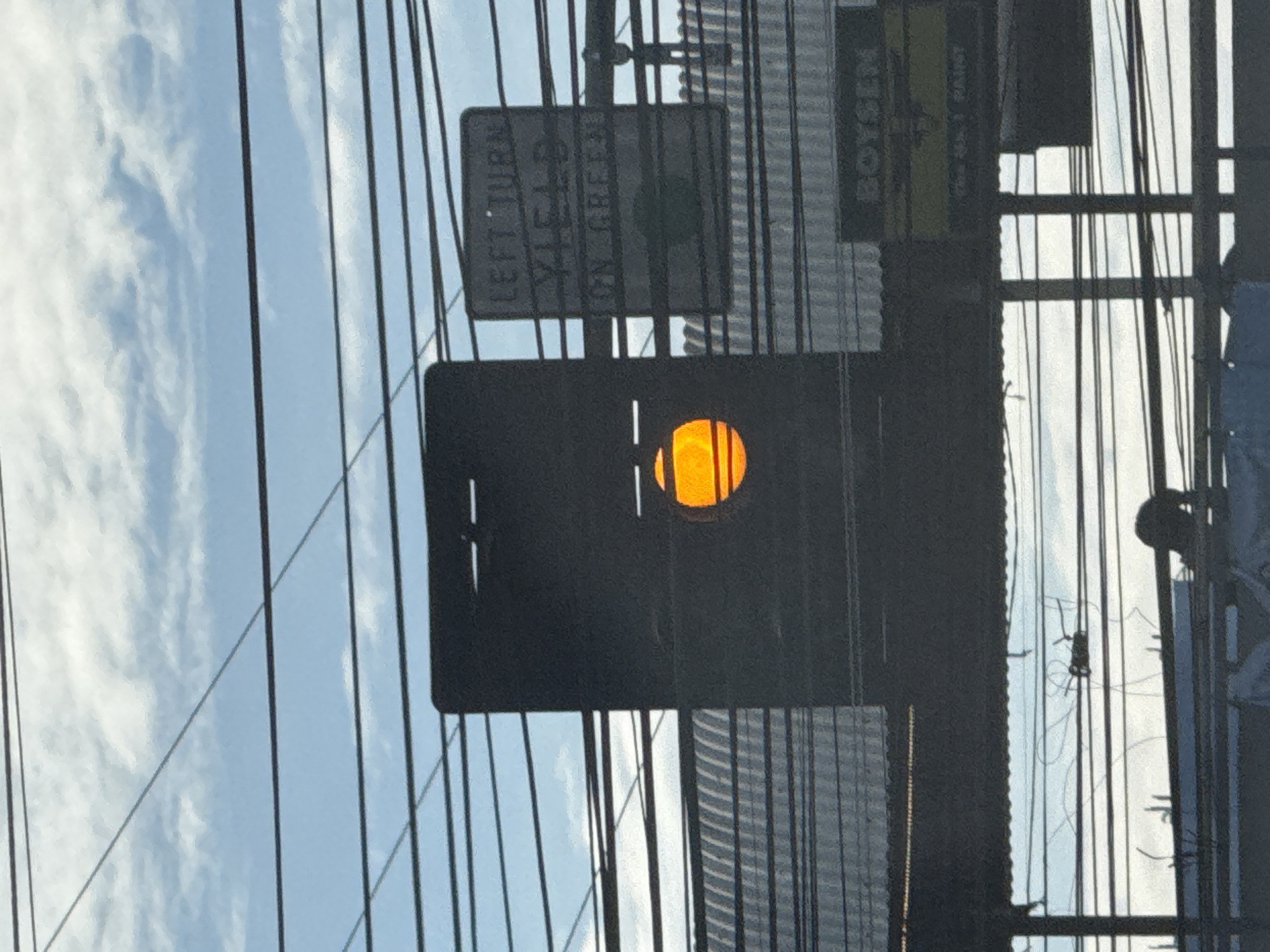 An outdoor shot captures a traffic light displaying an amber signal, positioned amidst a web of electrical wires against a backdrop of a cloudy blue sky. To the right of the traffic light, a sign reads "LEFT TURN YIELD ON GREEN." Below the traffic light and sign, a corrugated metal roof structure is visible, and further down, a sign displays the word "BOYSEN" in large lettering, with smaller text below indicating "QUALITY PAINT." The scene appears to be located along a street in Davao City, Philippines, where overhead electrical wires are a common feature. In the foreground, a blurry silhouette of a person can be seen, suggesting a street-level perspective.
