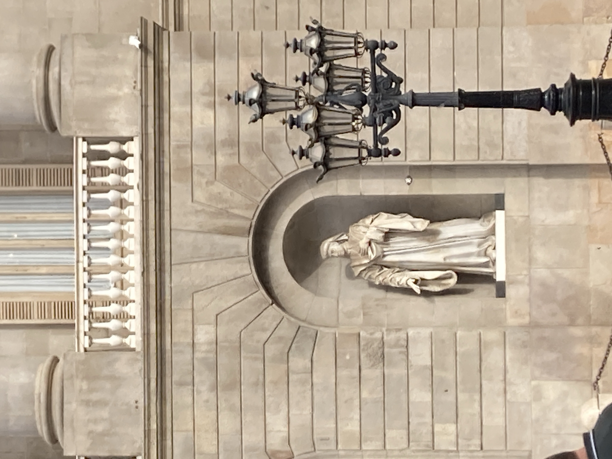 The media file shows a stone facade of a building, likely in Barcelona, Spain. The facade is made of large, rectangular stone blocks and features architectural elements such as columns, decorative moldings, and a semicircular niche. Within this niche, a pale stone statue of a robed figure is visible. The figure appears to be a religious or historical personage, depicted with a serene or contemplative expression.
Above and to the right of the niche, an ornate, dark metal lamppost with multiple lamps extends horizontally across the frame. The lamppost is a prominent object, suggesting it is part of the building's exterior or surrounding streetscape. To the left of the statue, a section of a balustrade with white spindles is visible, indicating a balcony or a similar elevated architectural feature. Further to the left, a glimpse of a window with horizontal blinds is also present.
The overall scene is static and architectural, with no obvious activity or event taking place. The lighting suggests it is daytime, and the weather appears to be clear, as there are no indications of rain or overcast skies. The style of architecture and the presence of such decorative elements are common in historic European cities like Barcelona. No visible text is present in the image.