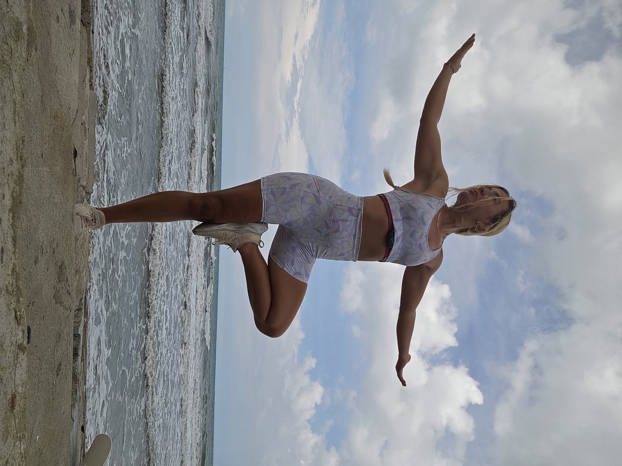 A woman with blonde hair is performing a yoga pose on a concrete platform overlooking the ocean. She is wearing a light-colored patterned sports bra and shorts, and light-colored athletic shoes. Her arms are outstretched to her sides, and her right leg is bent with her foot placed on her left thigh, resembling the Tree Pose. The ocean behind her has moderate waves, and the sky is filled with scattered white clouds. The foreground shows a rough, textured concrete surface. The scene appears to be a beach in Mũi Né, Vietnam, a popular coastal city known for its beaches and water sports.