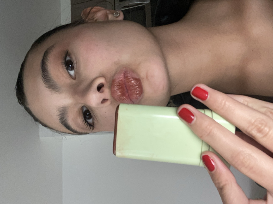 A person poses closely for a selfie with a puckered expression. Red nail polish adorns their fingernails. They are holding a rectangular, light green object close to their mouth. Their eyebrows are thick and prominent. An earring is visible on the left ear. The angle of the photo is tilted, and a cabinet is noticeable in the background.