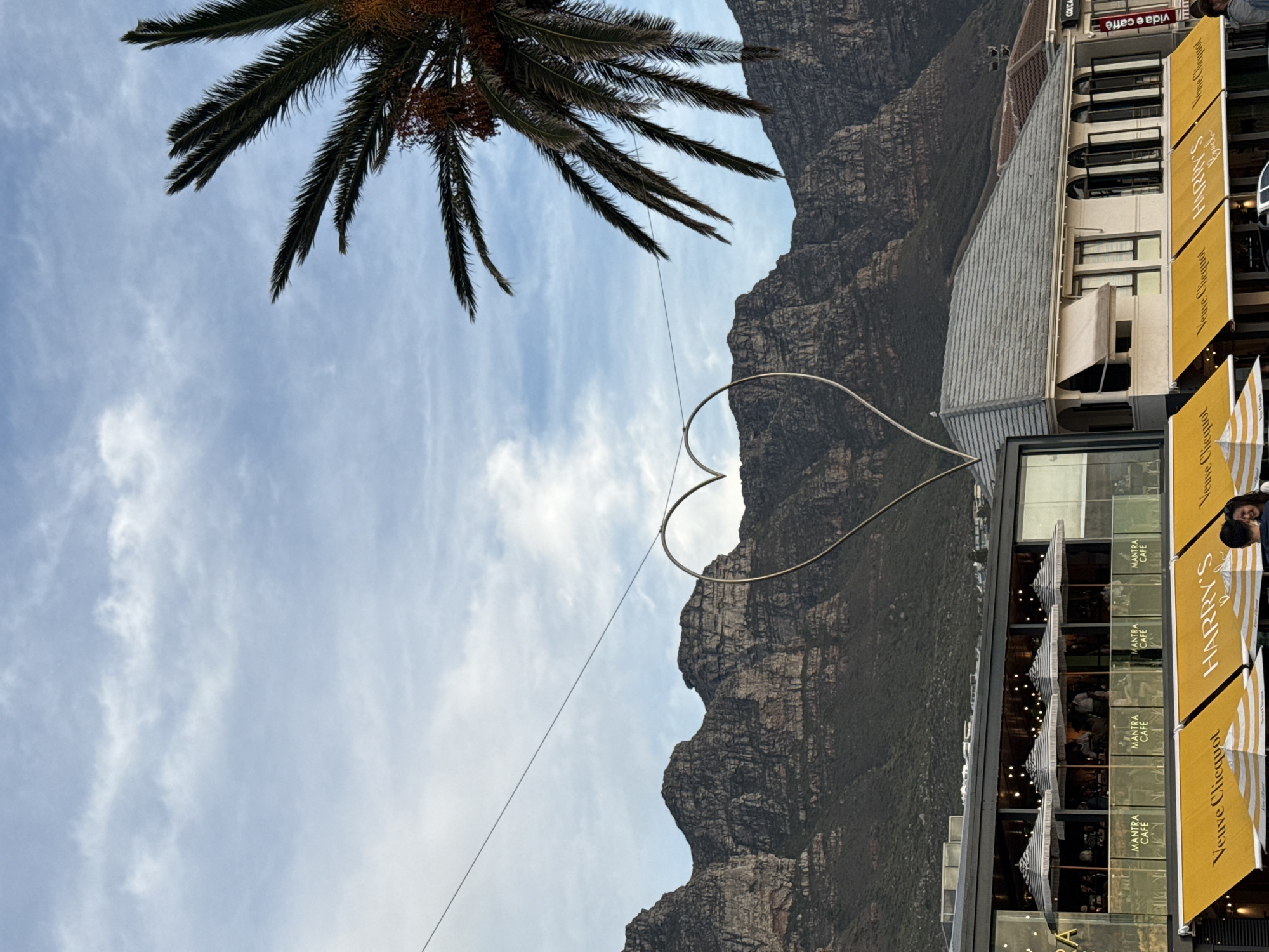 The image captures a scene in Cape Town, South Africa. The main subjects include Table Mountain, partially obscured by a palm tree, and a large metal heart structure suspended by wires in the foreground. In the lower right of the frame, there's a building featuring a cafe named "Mantra Cafe," with "Harry's" advertised alongside "Veuve Clicquot." People are visible inside the cafe through the windows. The sky is light blue with scattered clouds. The mountain is rocky and textured.