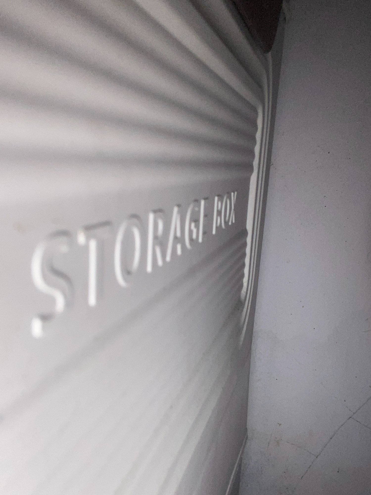 The image shows a close-up, angled view of the embossed text "STORAGE BOX" on the side of a white plastic storage container. The container has a ribbed texture. The background consists of a light-colored wall, which is slightly out of focus. The lighting creates shadows that highlight the texture of the container and the embossed letters. The image is taken indoors, likely in a residential or storage area. There are no people or any contextual elements that would indicate a specific location within Sepayung, Indonesia.