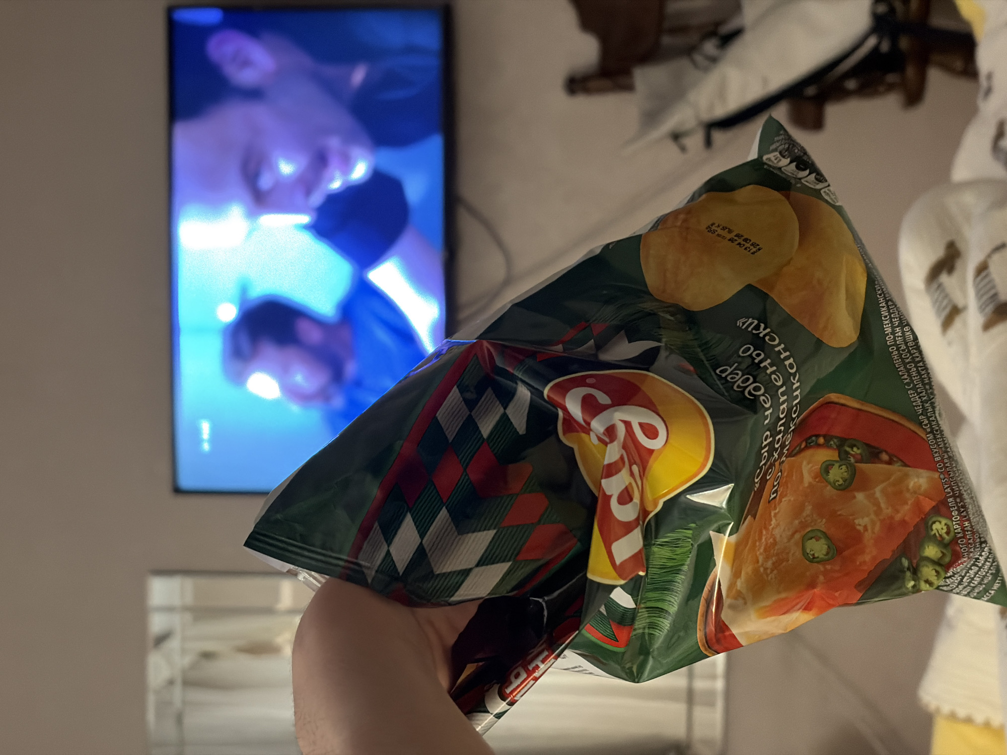The image depicts a casual indoor scene, likely in a living room or bedroom in Minsk, Belarus, during the evening or night. In the foreground, a hand holds a bag of Lay's potato chips. The bag is predominantly dark green with a red and white checkered pattern on one side, and features the Lay's logo along with illustrations of golden potato chips and a savory dish (resembling a pizza or nachos) garnished with jalapeño slices.
In the background, a flat-screen television is mounted on a wall, displaying a blurry image of at least two individuals, possibly from a movie or TV show. The figures on the screen are illuminated by a distinct blue and purple light. Below the TV, a reflective surface, possibly a table or shelf, is visible.
Visible text on the chip bag, predominantly in Cyrillic, identifies the flavor as "Сыр чеддер по-мексикански" (Cheddar cheese Mexican-style) and "халапеньо" (jalapeño). A production code "13 04 28SR S96092" is also discernible on one of the illustrated chips. The scene suggests a relaxed moment of watching television and snacking.