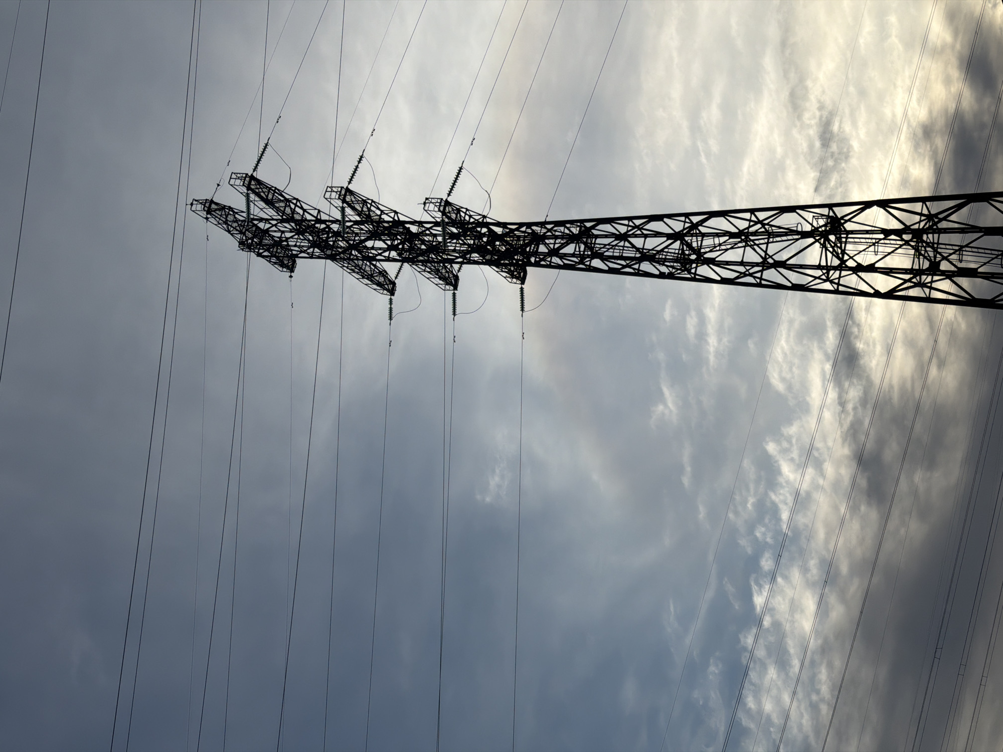 A metal electrical transmission tower stands against a backdrop of cloudy sky. The tower is composed of a lattice-like network of metal beams and crossbars, its structure appearing complex and angular. High-voltage power lines, which are suspended from the tower via insulators, stretch across the scene. The tower dominates the view and is shown in detail, while the sky in the background has a cloudscape with varying shades of gray and white.
It is not possible to verify the location in the city of Chaikovsky, Russia from the image alone.
