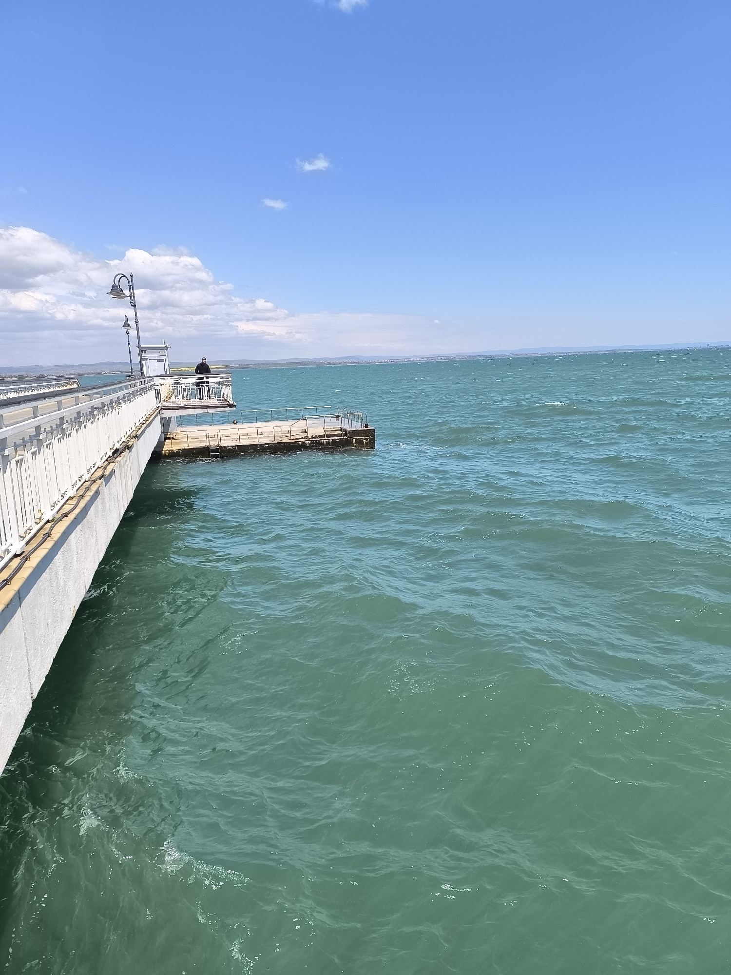 The image shows a concrete pier extending into a body of water, likely the Black Sea. The pier has a white railing and a lamppost. A small platform extends off the pier, near a figure standing on the main pier. The water is greenish-blue, and the sky is blue with scattered clouds. The skyline in the distance suggests a city, possibly Burgas, Bulgaria.