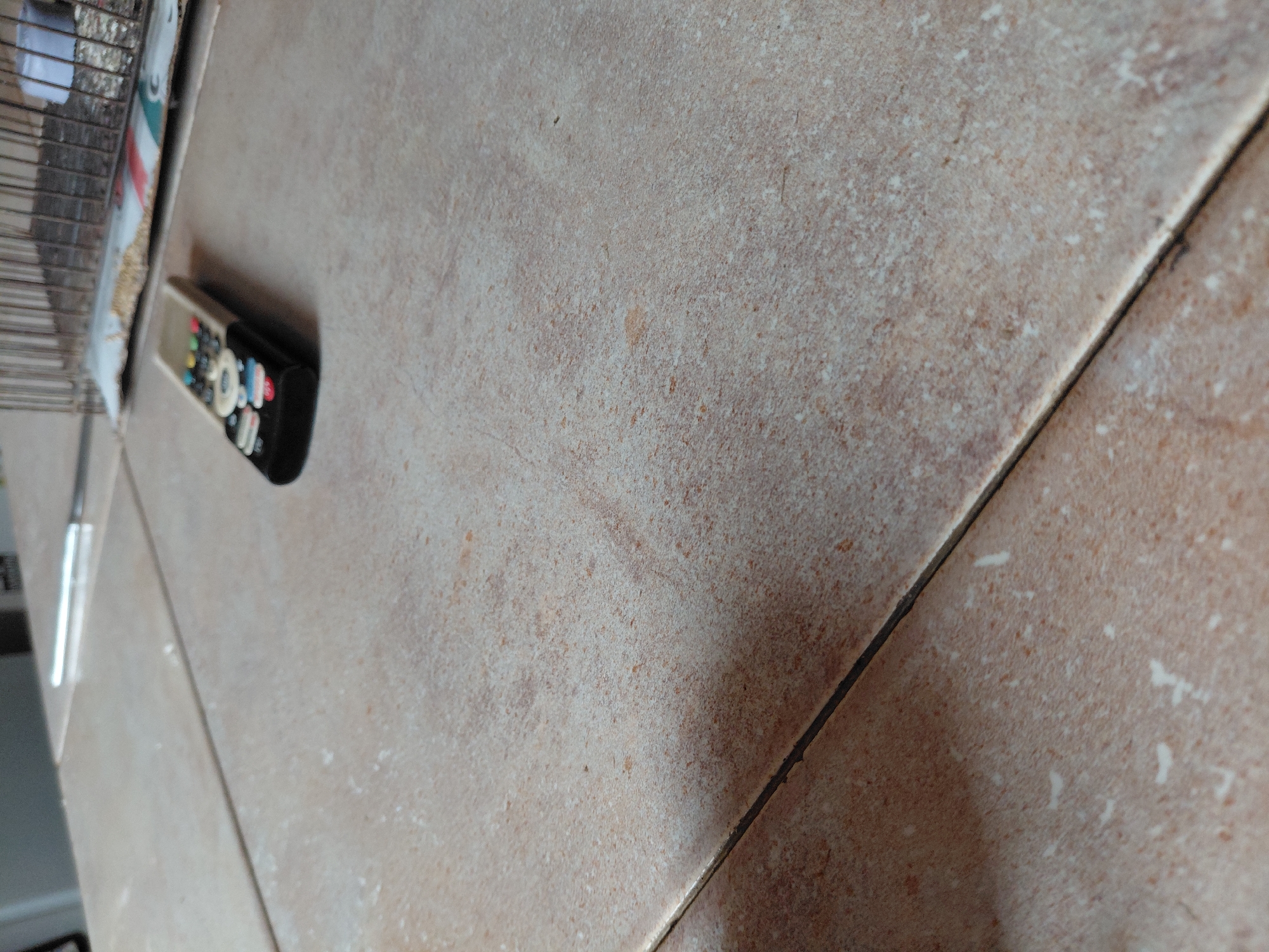 The image displays a close-up, angled view of a tiled surface in an indoor setting, likely in Santa Ana, Venezuela. The tiles are light beige or sandy brown with a subtle, granular texture and varying speckles across their surface. Dark grout lines demarcate the individual tiles, running diagonally across the frame.
Prominently featured on the left side, resting on the tiled surface, is a modern remote control. It has a black base and a lighter-colored top, with an array of small, colorful buttons suggesting dedicated functions or streaming service shortcuts, typical of smart TV remotes. While specific text on the buttons is not legible due to focus and size, their distinct colors and placement are visible.
To the far left, a portion of a metal grid or cage structure is partially visible and out of focus, possibly belonging to a household appliance or storage unit. A thin, reflective strip, which could be a metal edge or a ruler, also appears near the upper left corner.
No human subjects are present, and no activity or event is taking place; the objects are static. The lighting is consistent with an indoor environment, though the specific time of day is not evident. No clear, decipherable text is visible beyond the general impression of icons on the remote's buttons. The scene conveys a simple, everyday domestic or commercial environment.
