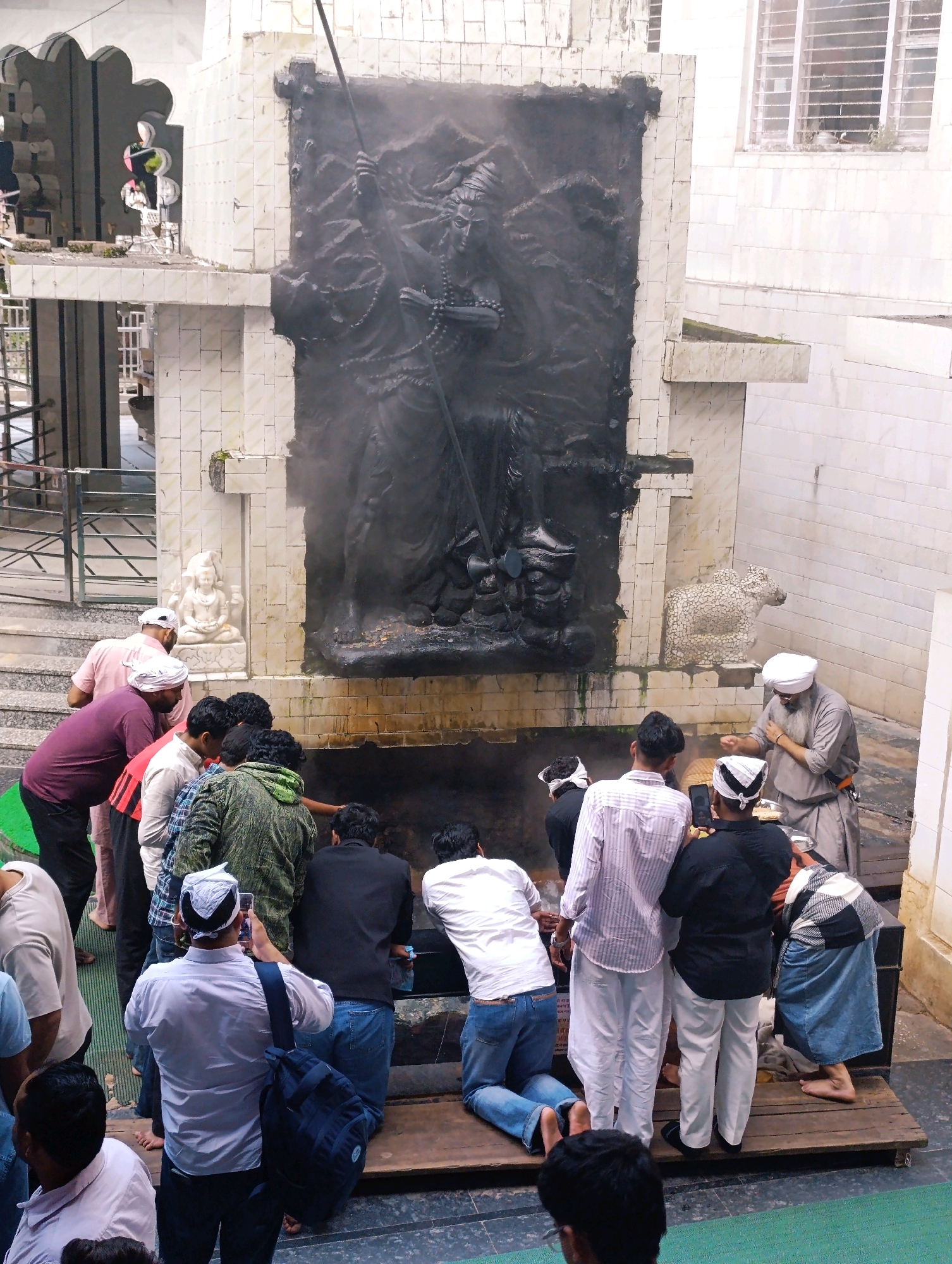 In Kasol, India, a group of individuals, many of whom appear to be men, are gathered around a large, dark relief sculpture. The sculpture depicts a figure with a staff, possibly religious in nature. Some individuals are kneeling, while others stand, observing the sculpture. One person is taking a photograph with a mobile phone. The environment appears to be an outdoor religious site, with tiled walls and structures. A small statue of a seated figure is visible to the left of the main sculpture. To the right, a stone carving of a bull-like creature is present. Smoke or mist is emanating from the base of the main sculpture, adding a dramatic element to the scene. The ground in front of the sculpture is a dark, wet surface.
