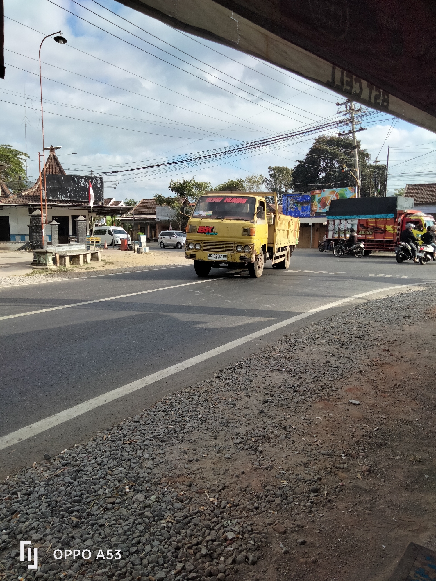 The image captures a street scene in Wonoanti, Indonesia, on a sunny day with a cloudy sky. A yellow truck, bearing the license plate "AG 8290 RN," is driving on the asphalt road, which is marked with white lines. To the left of the road, there is a building with a red and white flag and a street lamp. Several motorcycles are also visible on the road. In the background, there are more buildings and trees. The foreground of the image shows a gravelly shoulder of the road. The bottom left corner of the image displays the text "OPPO A53," indicating the device used to capture the photo.
