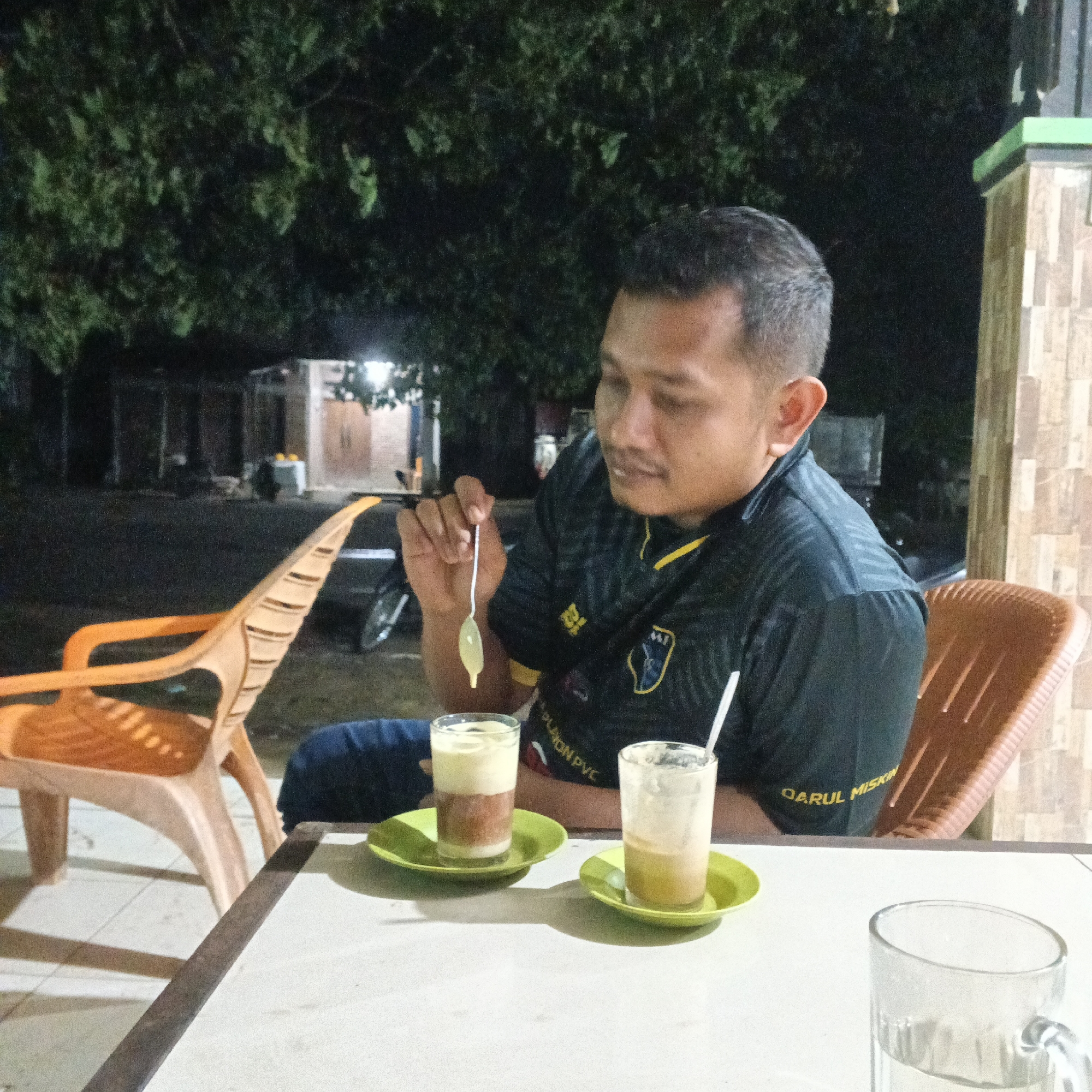 A man with short dark hair and a mustache, wearing a black t-shirt with yellow accents and a logo, is sitting at a table outdoors at night. He is holding a spoon with a green substance on it, which he appears to be tasting. In front of him are two glasses of layered drinks on green saucers, and a third empty glass. The background shows trees and some buildings, with lights on. To his left is an orange plastic chair, and to his right is a brown plastic chair. The table is white.