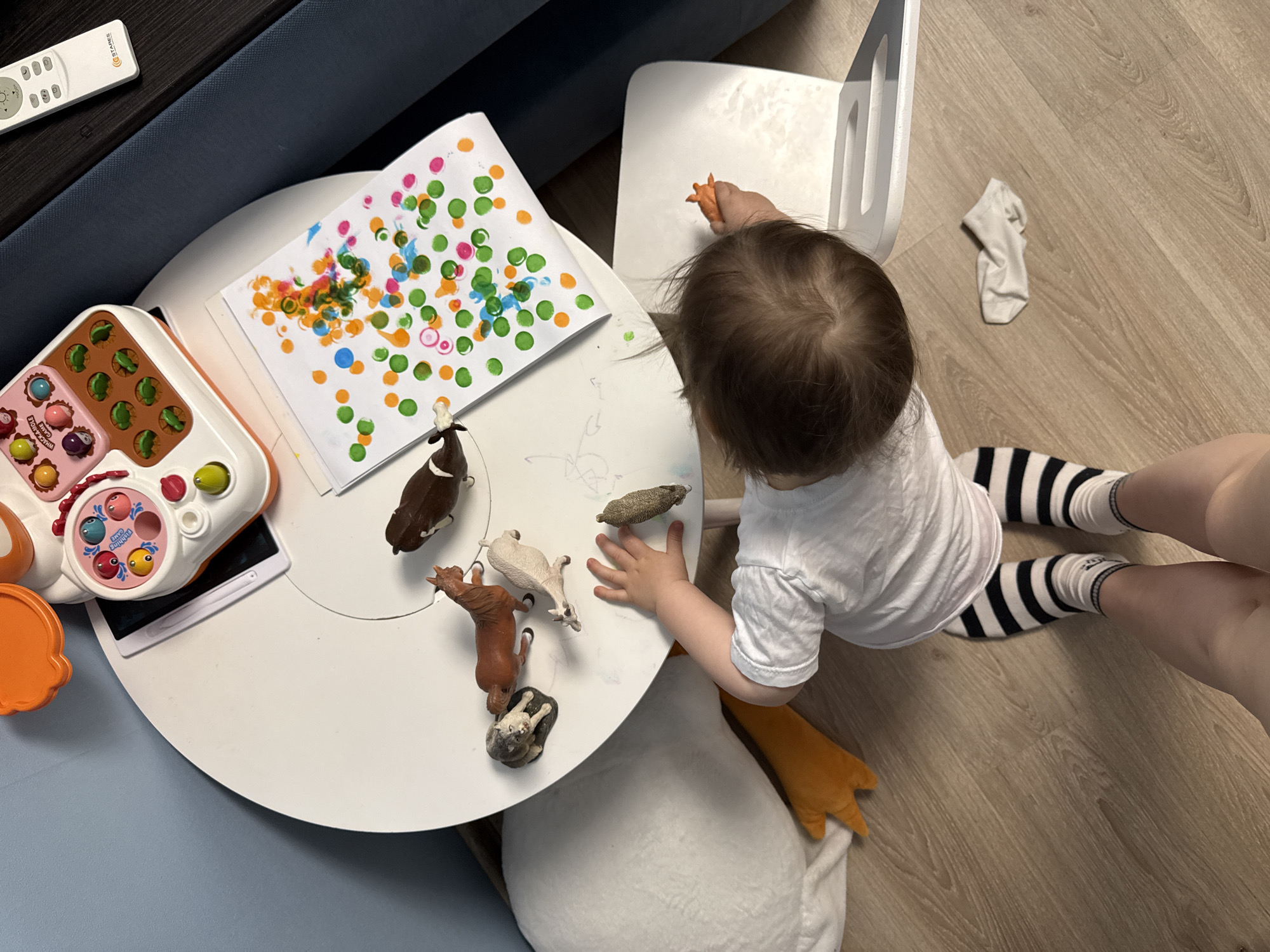 A high-angle shot depicts a child playing on a round white table. The child, wearing a white shirt and black-and-white striped socks, sits on the floor with an orange stuffed animal. The table holds a child's learning toy, a piece of paper covered in colored dots, and several toy animals. A small, white child's chair is nearby. A sock lies on the wooden floor. A sofa and a remote control are visible in the background. There is no evidence that this image was taken in Moscow, Russia.